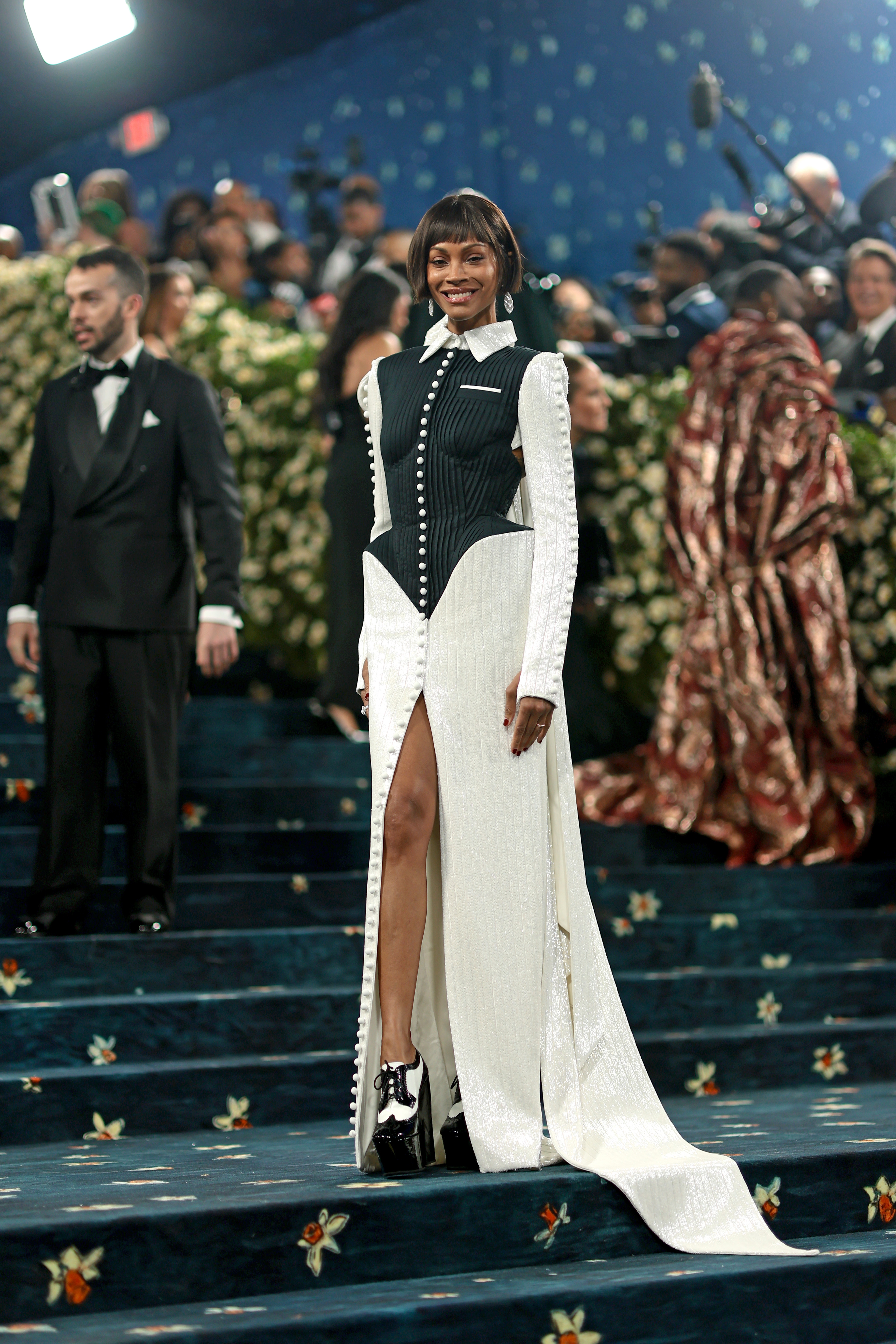 Person on a carpeted stairway wearing a stylish long dress with high slit and unique design elements, surrounded by people and photographers