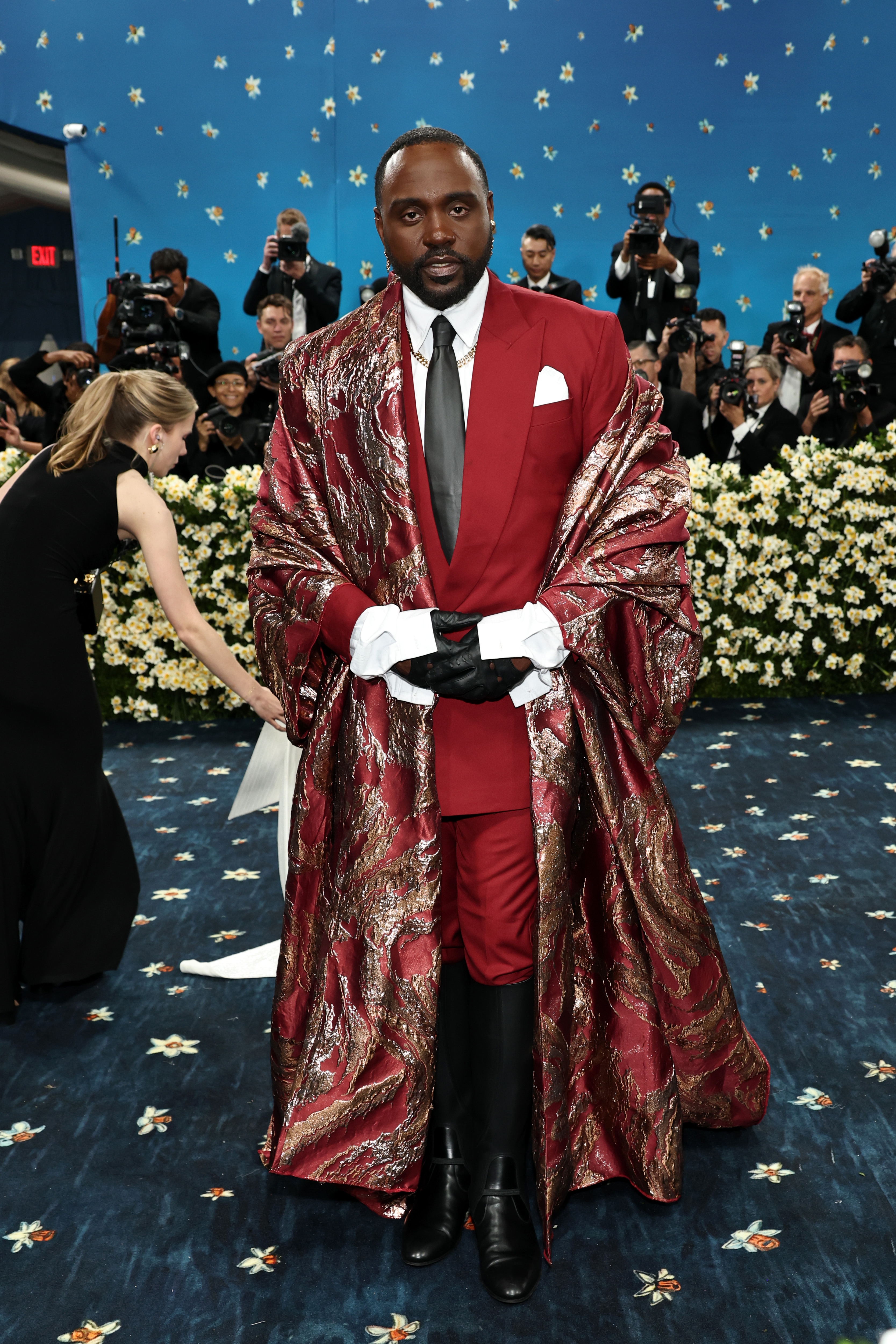 Person in a formal suit with an ornate cape on a red carpet, surrounded by photographers