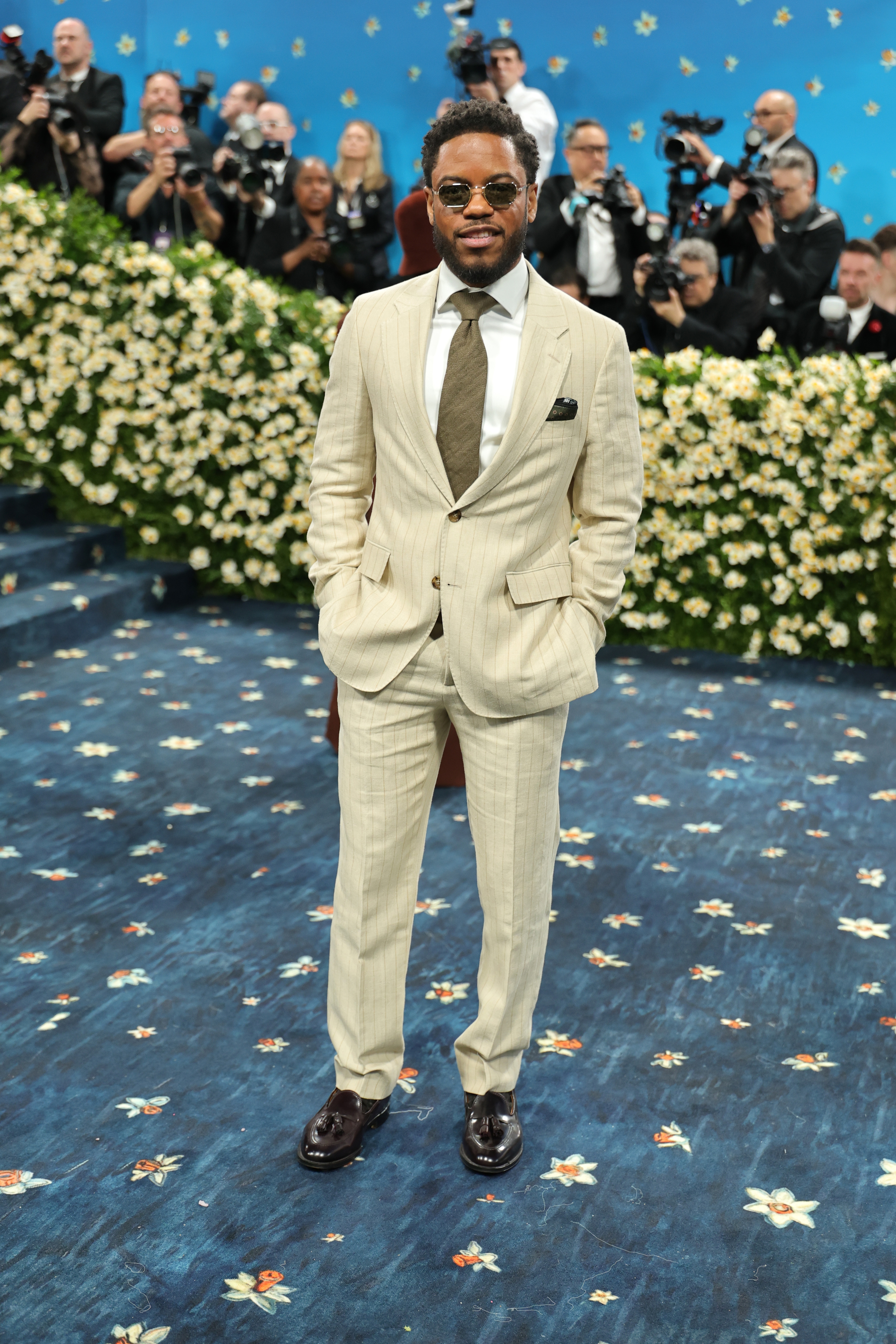 Man in tailored suit and tie on a floral-patterned carpet, surrounded by photographers at a celebrity event