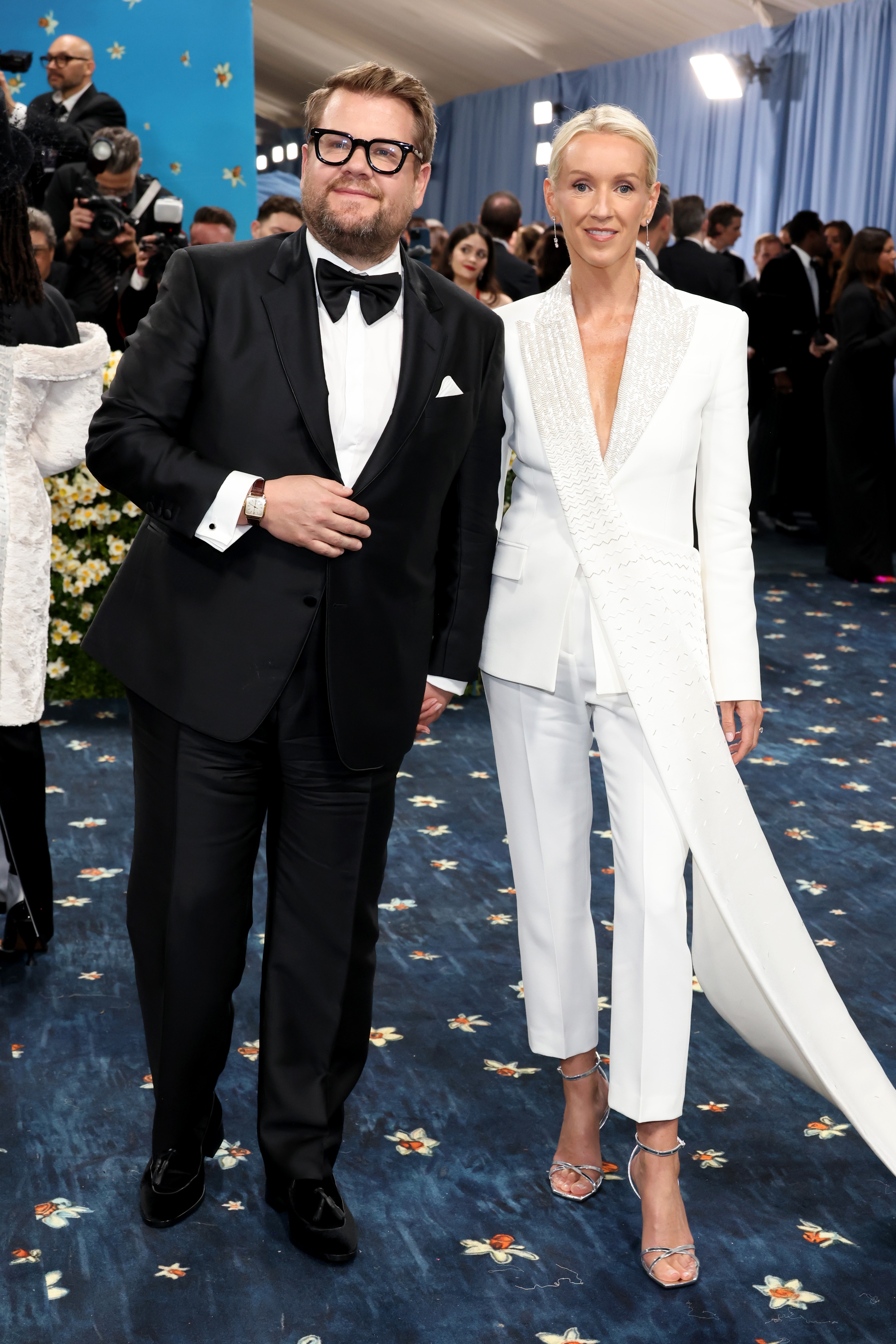 Man in tuxedo and woman in elegant white suit on event blue carpet, surrounded by attendees and photographers in background