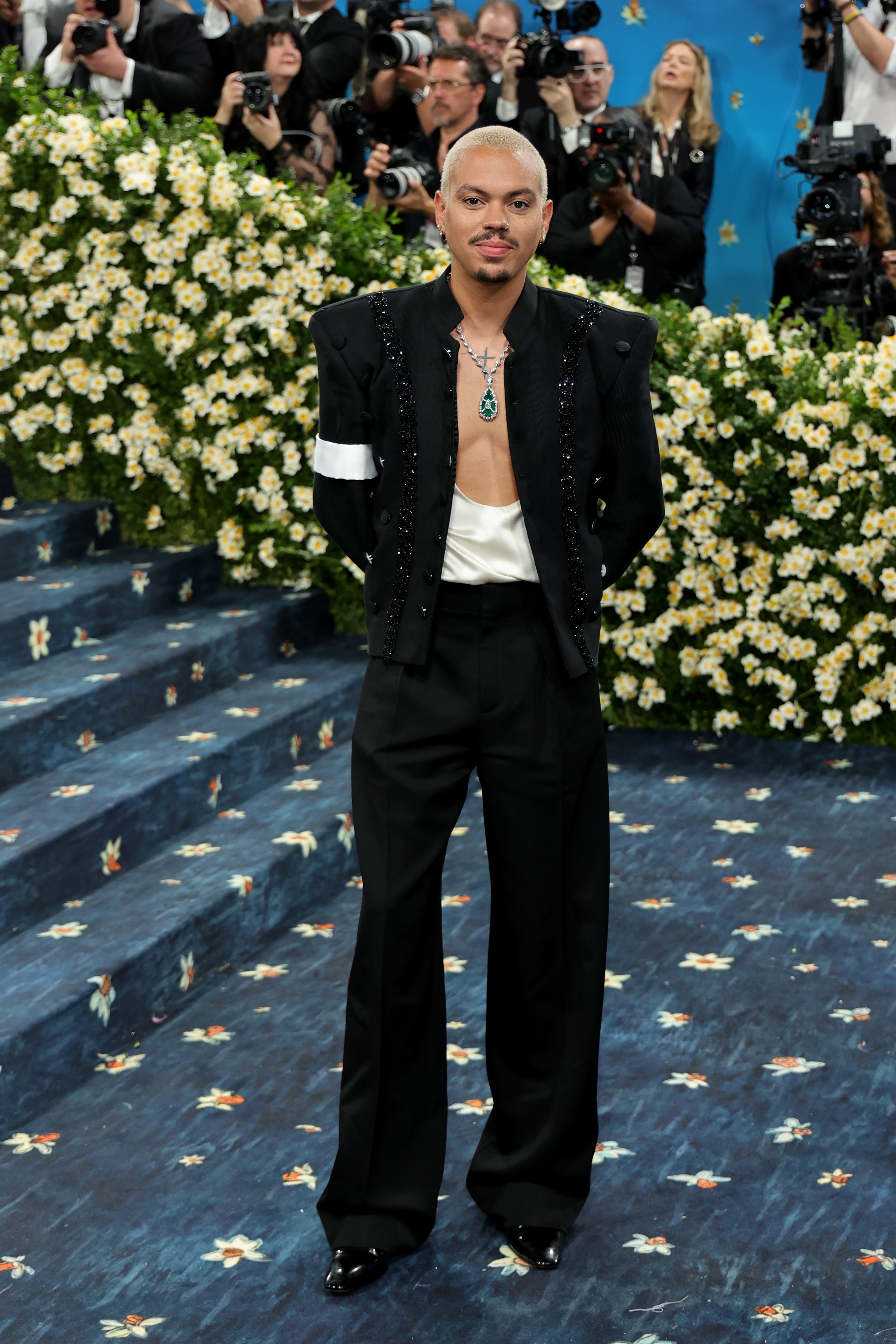 Person in a black embellished suit with a white armband and open shirt on a floral-patterned carpet at a celebrity event, surrounded by photographers
