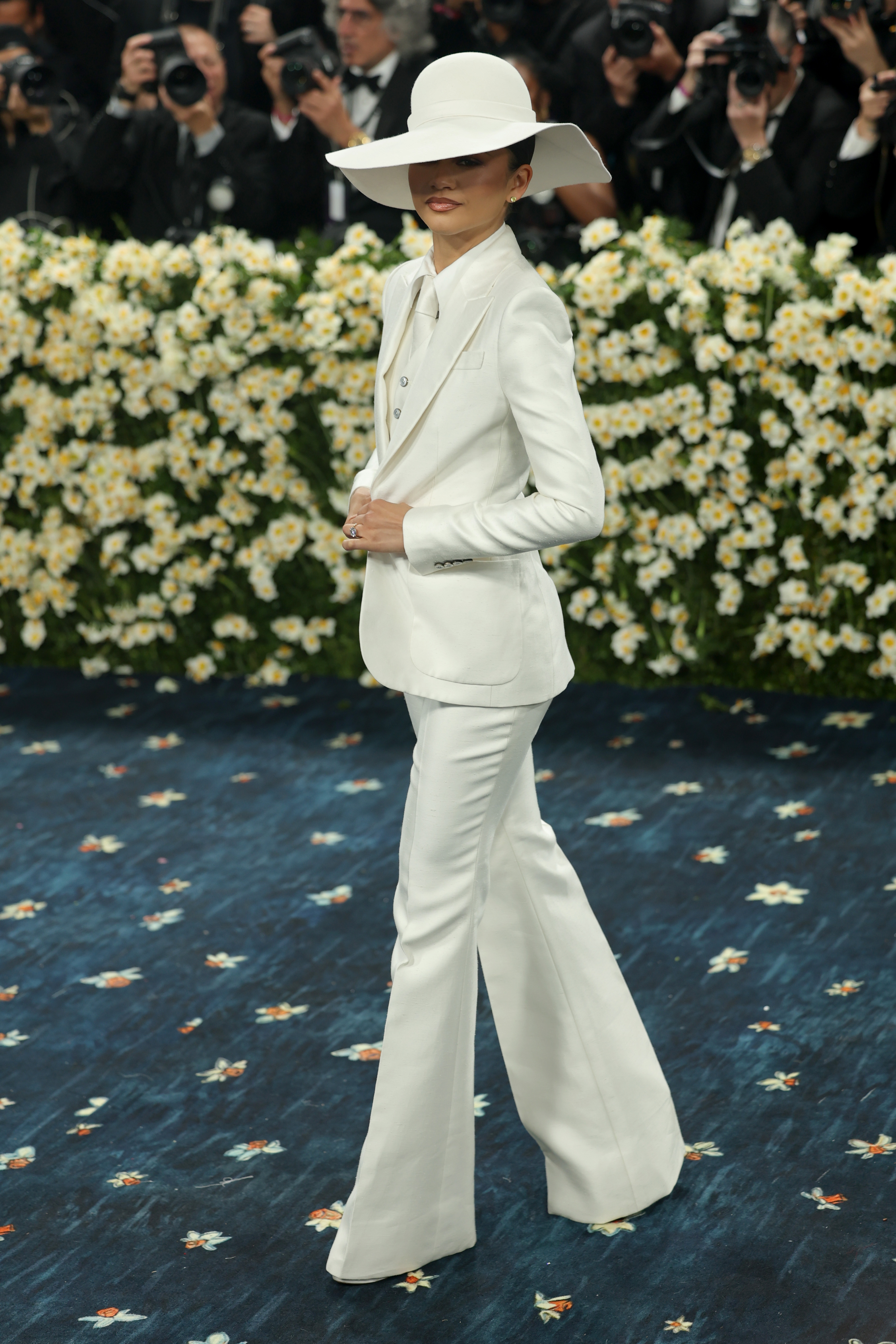 Person in a stylish white suit and wide-brimmed hat posing on a flower-patterned carpet while photographers capture the moment