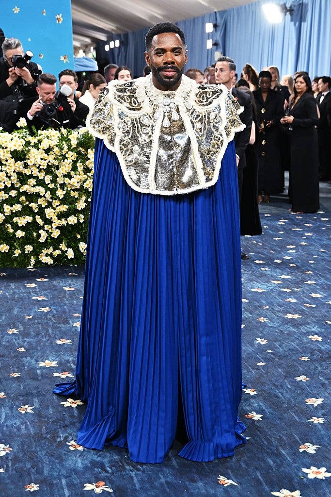 Person wearing an ornate, embellished cape and gown on a formal event carpet, surrounded by photographers and attendees