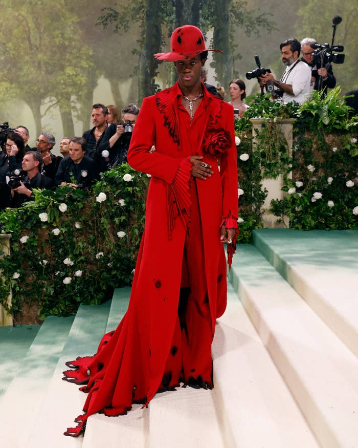 Person in elegant, long red coat and hat with black accents, posing on steps amidst paparazzi and greenery at a high-profile event