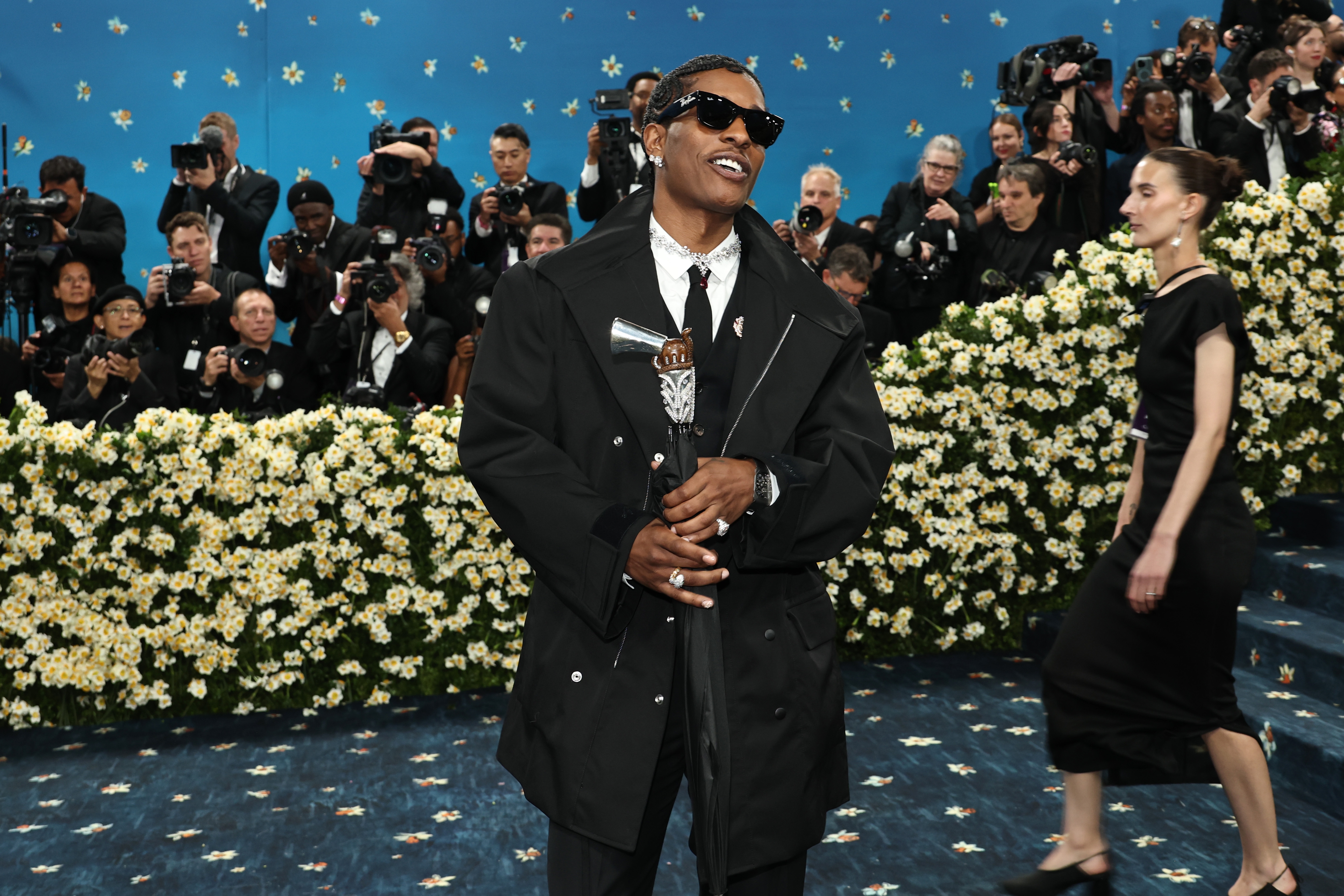 A$AP Rocky in oversized jacket and sunglasses poses on floral-decorated carpet while photographers take pictures