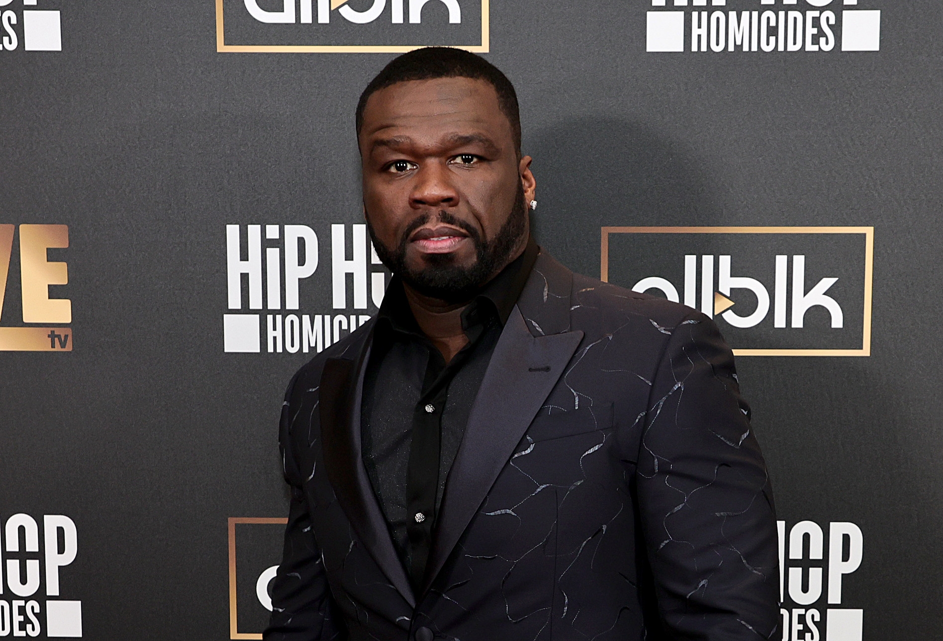 A person in a stylish patterned suit jacket poses at a &quot;Hip Hop Homicides&quot; event backdrop