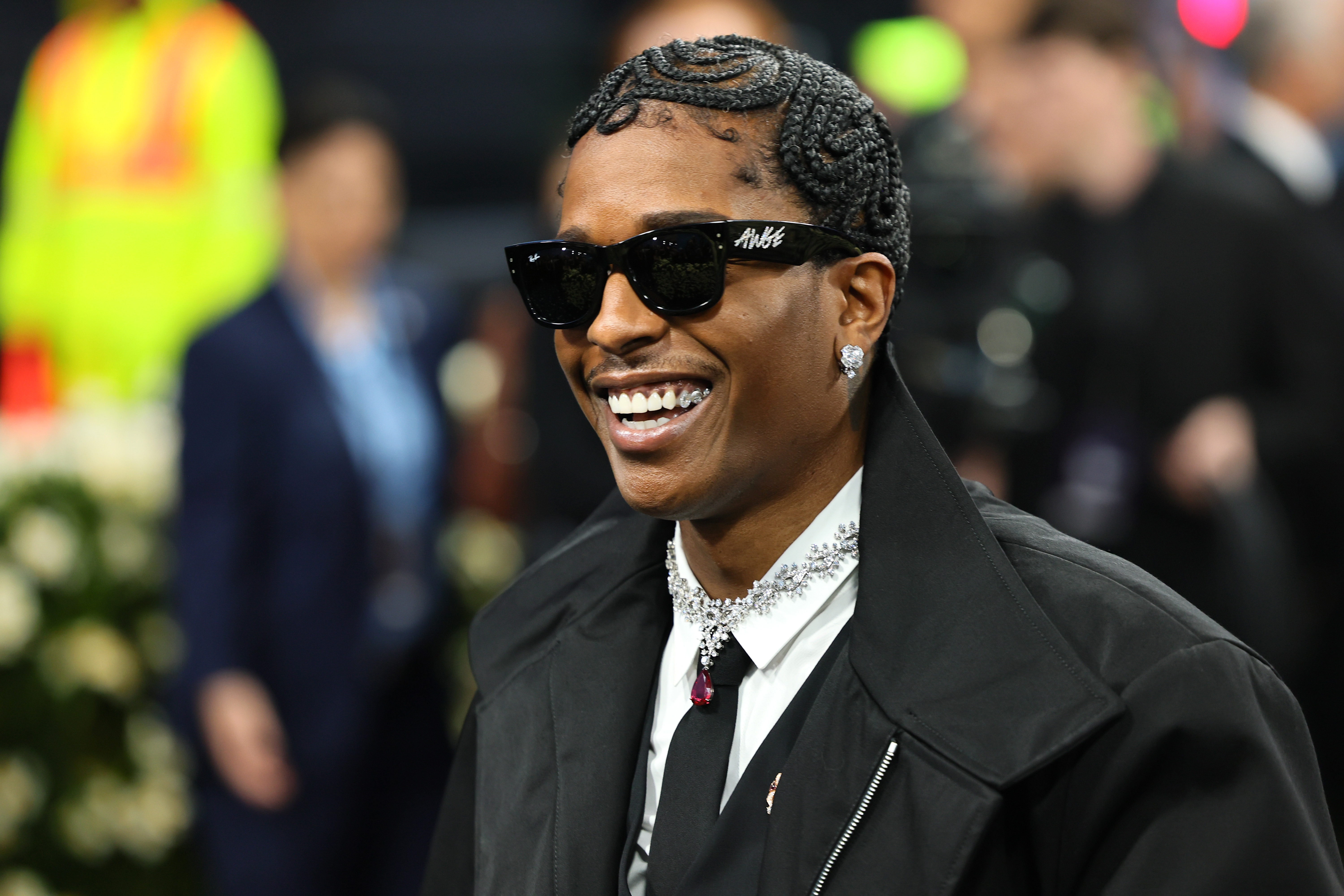 A$AP Rocky in formal attire with ornate hair twists, wearing sunglasses and a jeweled necklace, smiling at an event