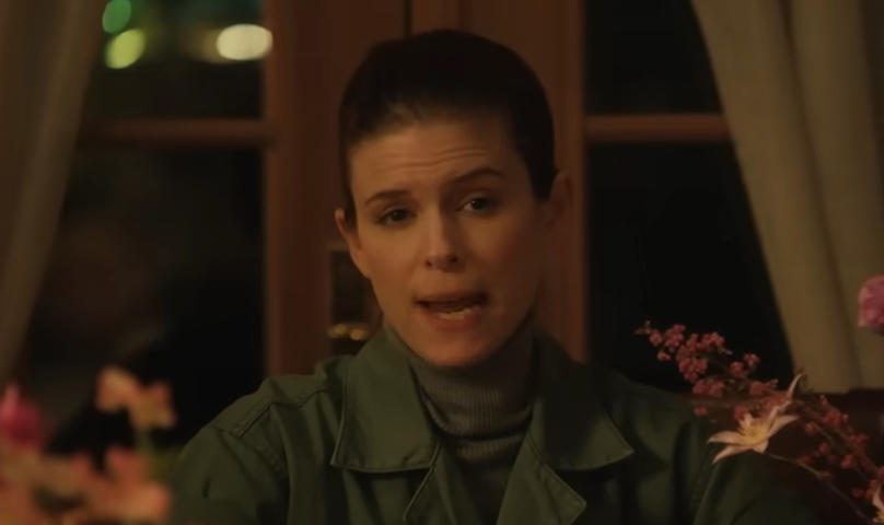 A person sitting at a table surrounded by flowers, wearing a green shirt and gray turtleneck, appears to be speaking