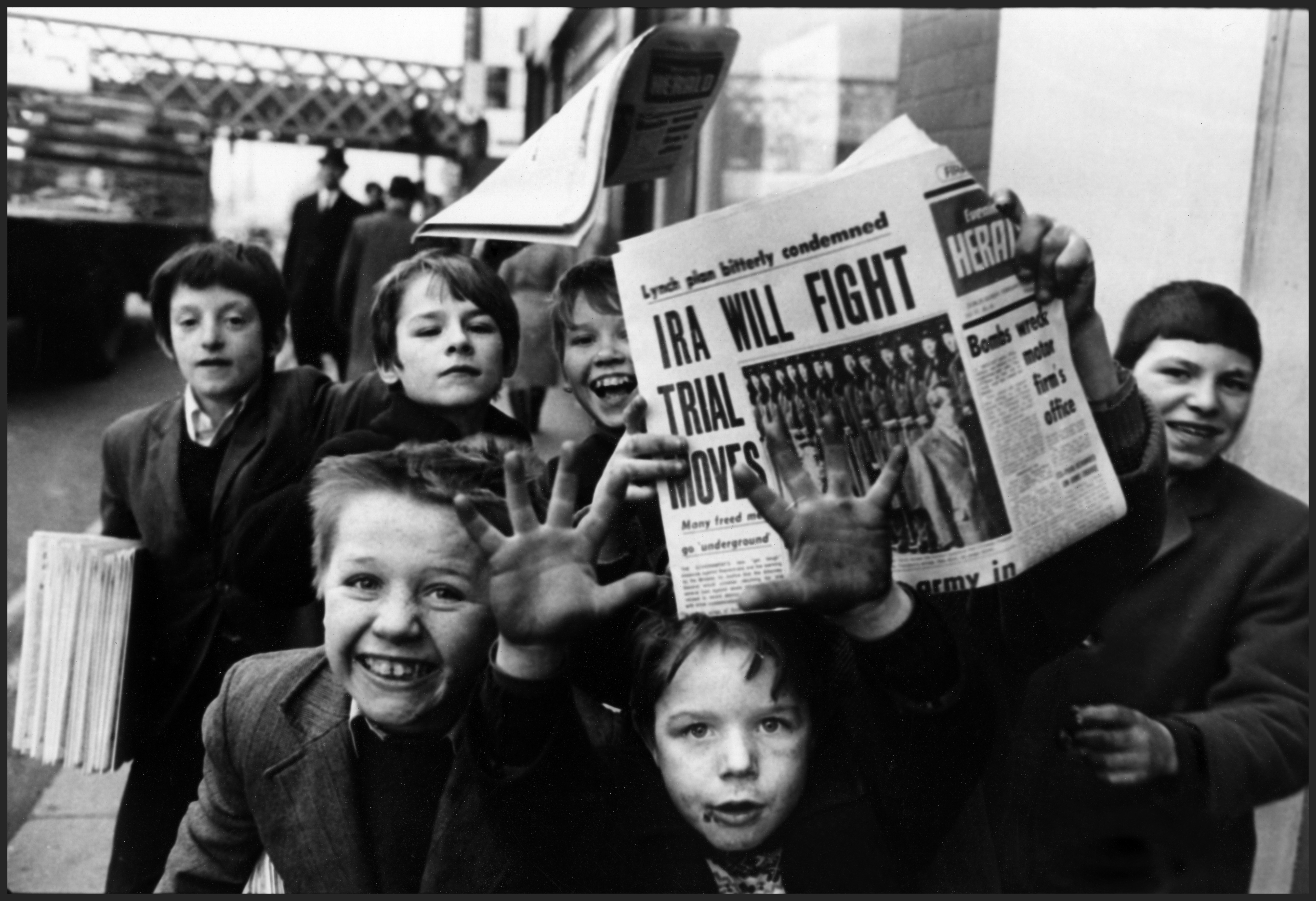 Group of children excitedly hold up newspapers with headline "IRA Will Fight Trial Moves" on a street