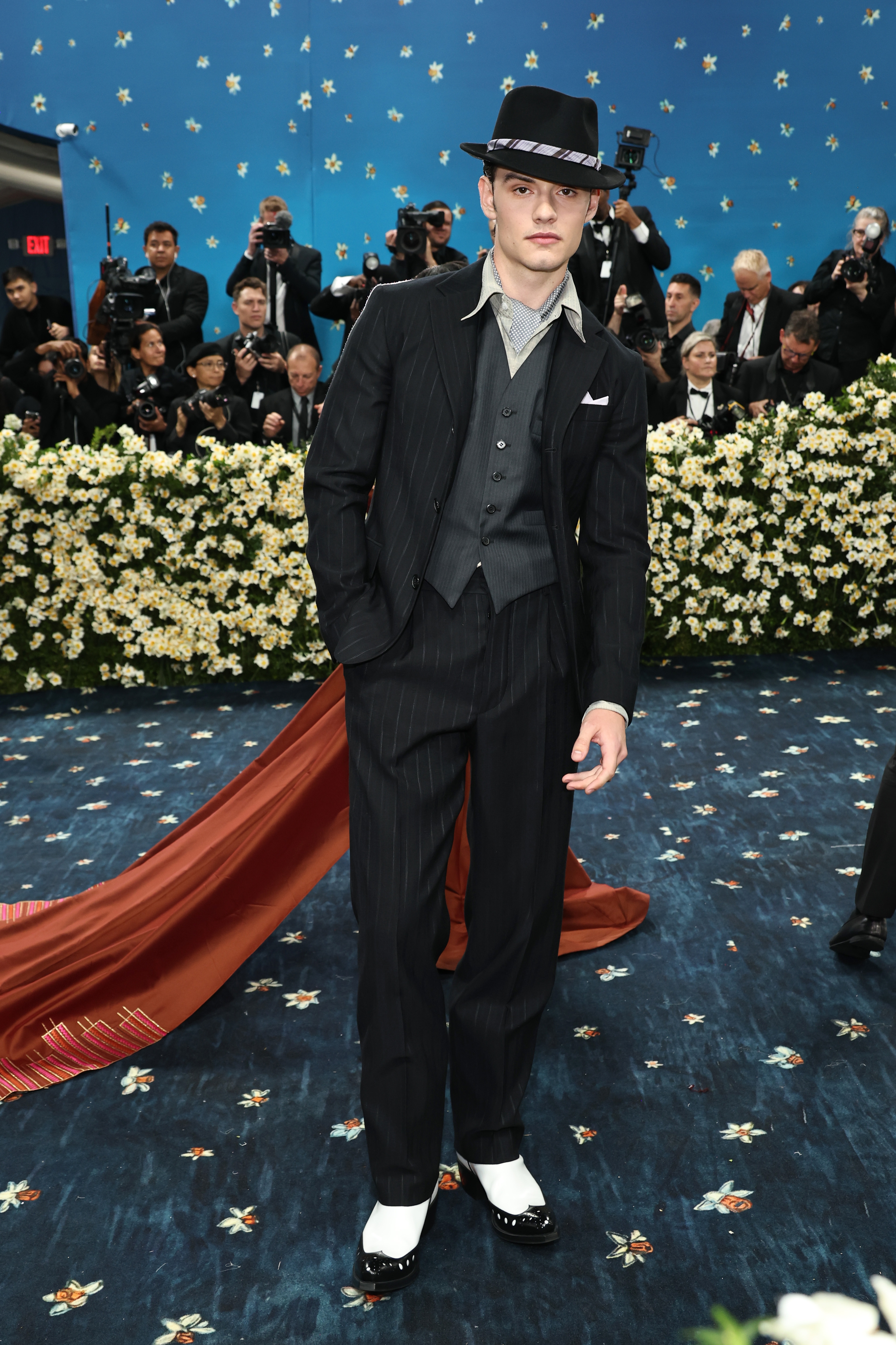 Person in a pinstripe suit, vest, and fedora on a floral carpet, surrounded by photographers