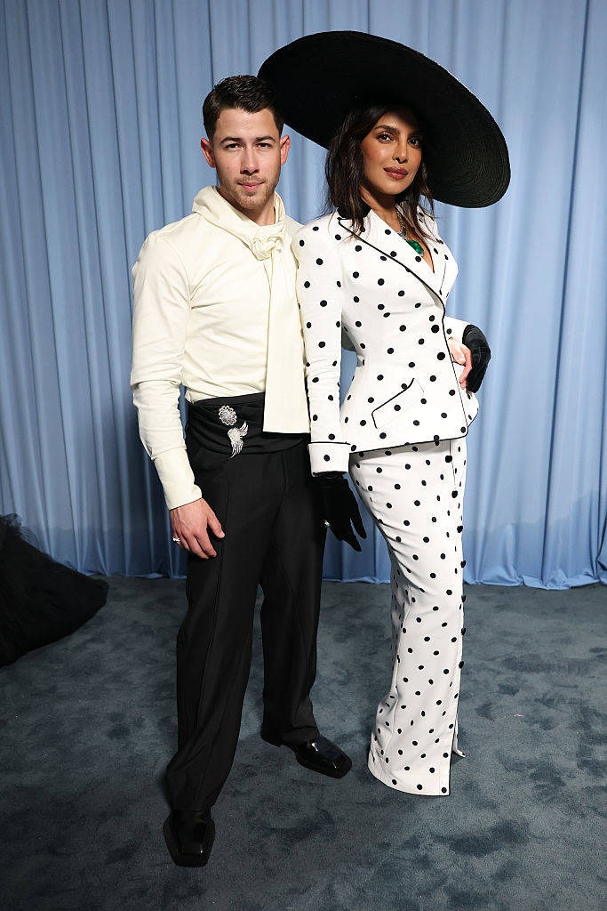 NIck in a shirt with a unique design and dark pants; Priyanka wears a polka-dot suit and large hat