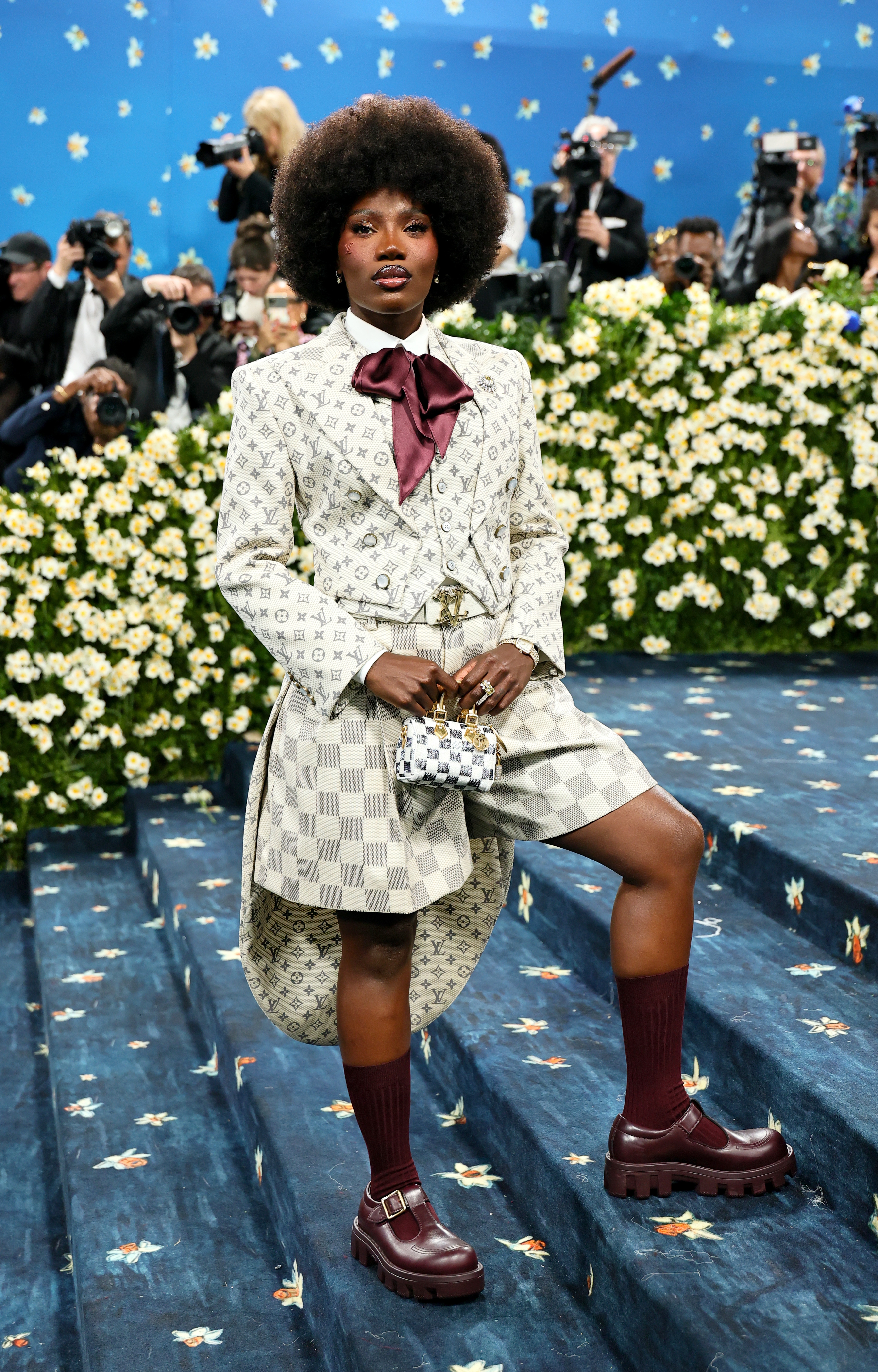Person wearing a patterned suit and shorts with bold platform shoes and a large bow, posing on a floral-themed red carpet