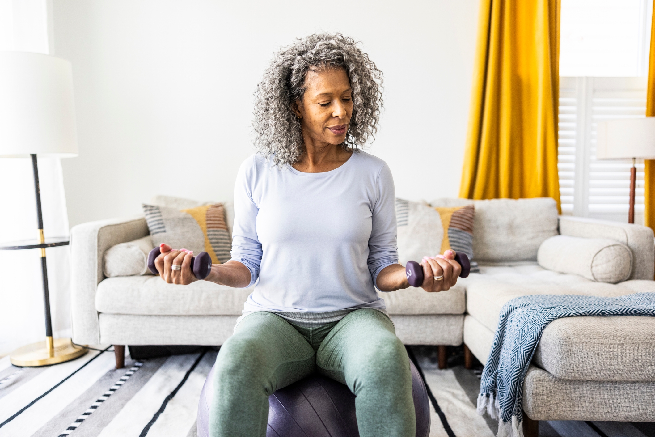 A woman sits on an exercise ball in a living room, lifting small dumbbells in both hands during a workout