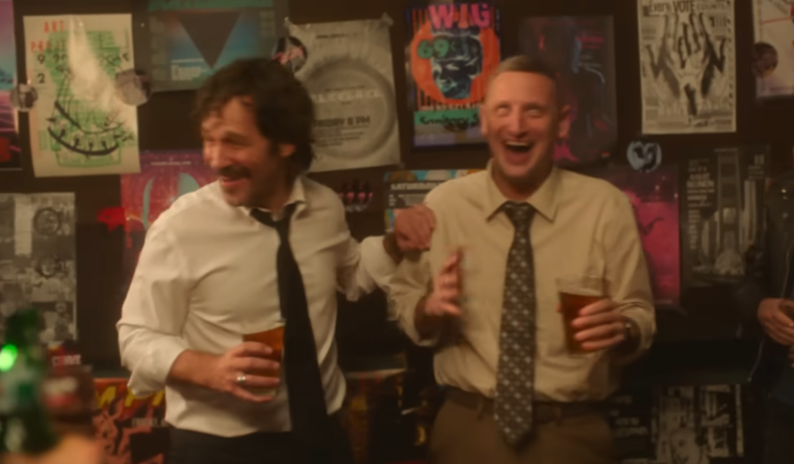 Two men in a casual setting, laughing and holding drinks, with posters in the background. One is in a tie and shirt, the other in a shirt and trousers
