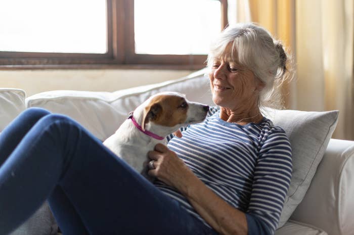Older woman in striped shirt smiling at a small dog on her lap while sitting on a couch