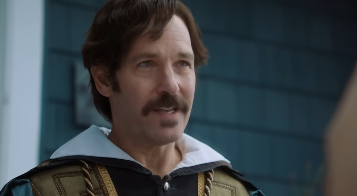 A person in period costume with a mustache speaks outdoors, wearing a chainmail design and a white collar