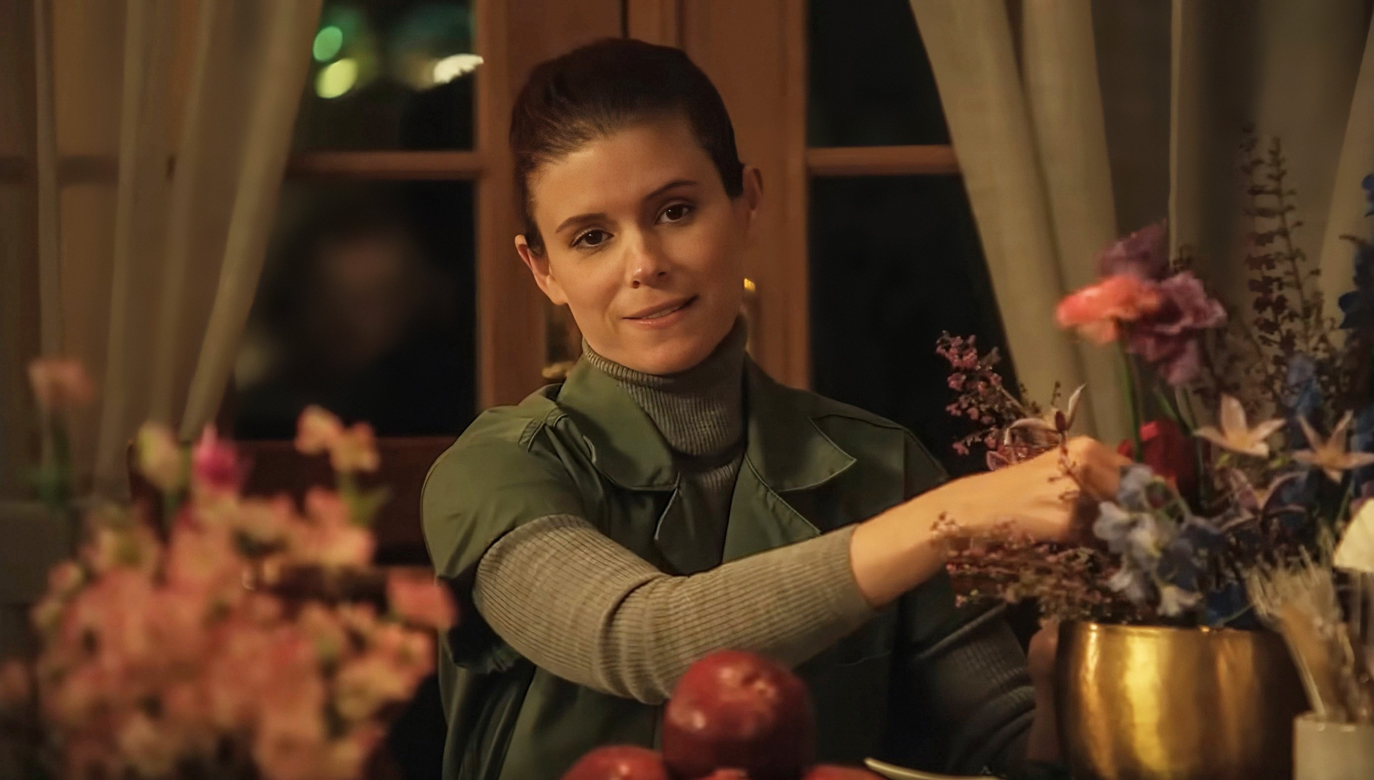 A person arranges flowers at a table with a calm expression. They wear a green apron over a turtleneck, surrounded by floral arrangements and fruits