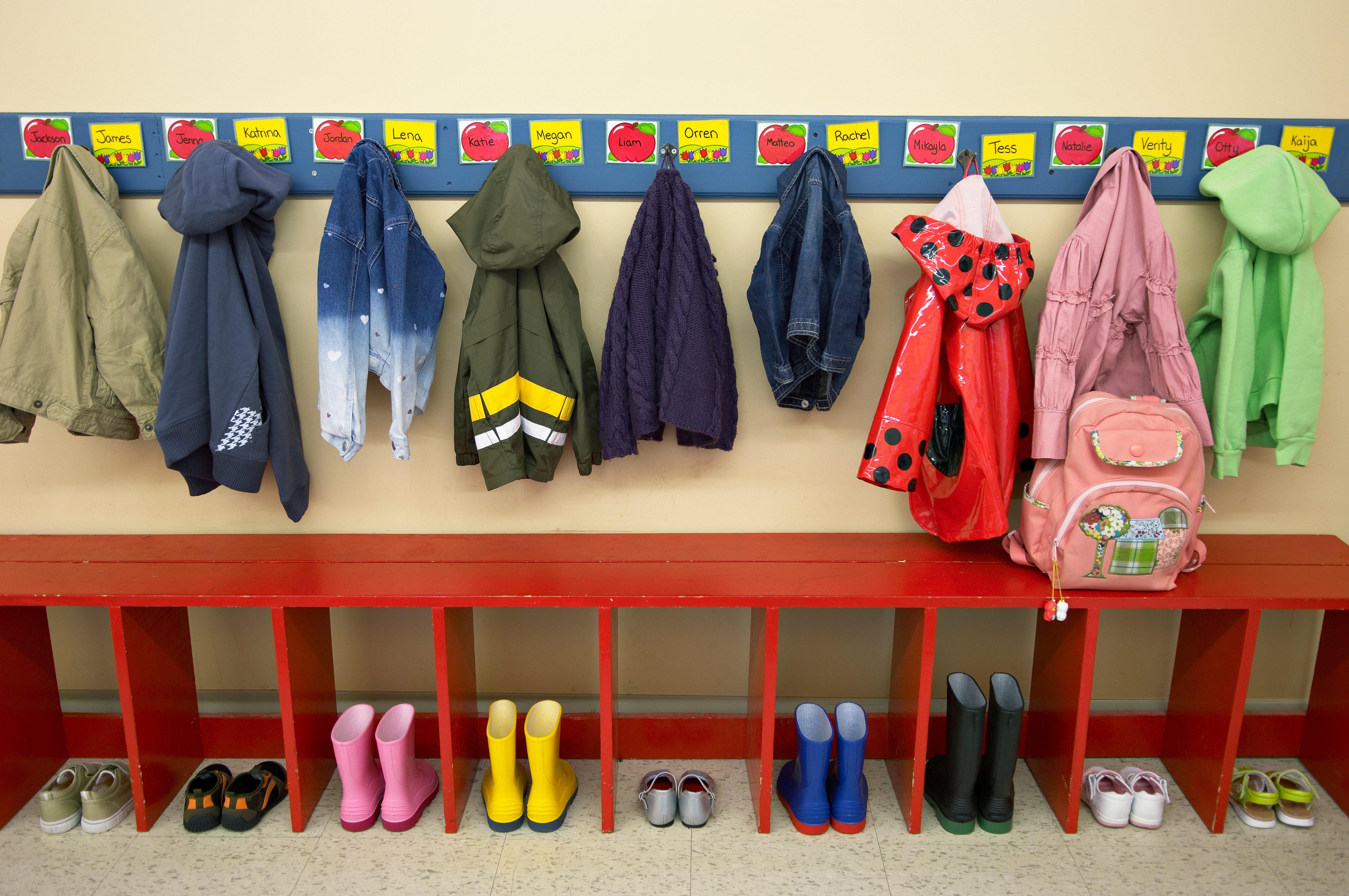 Children's jackets and backpacks hang on hooks above cubbies with rain boots below in a classroom setting