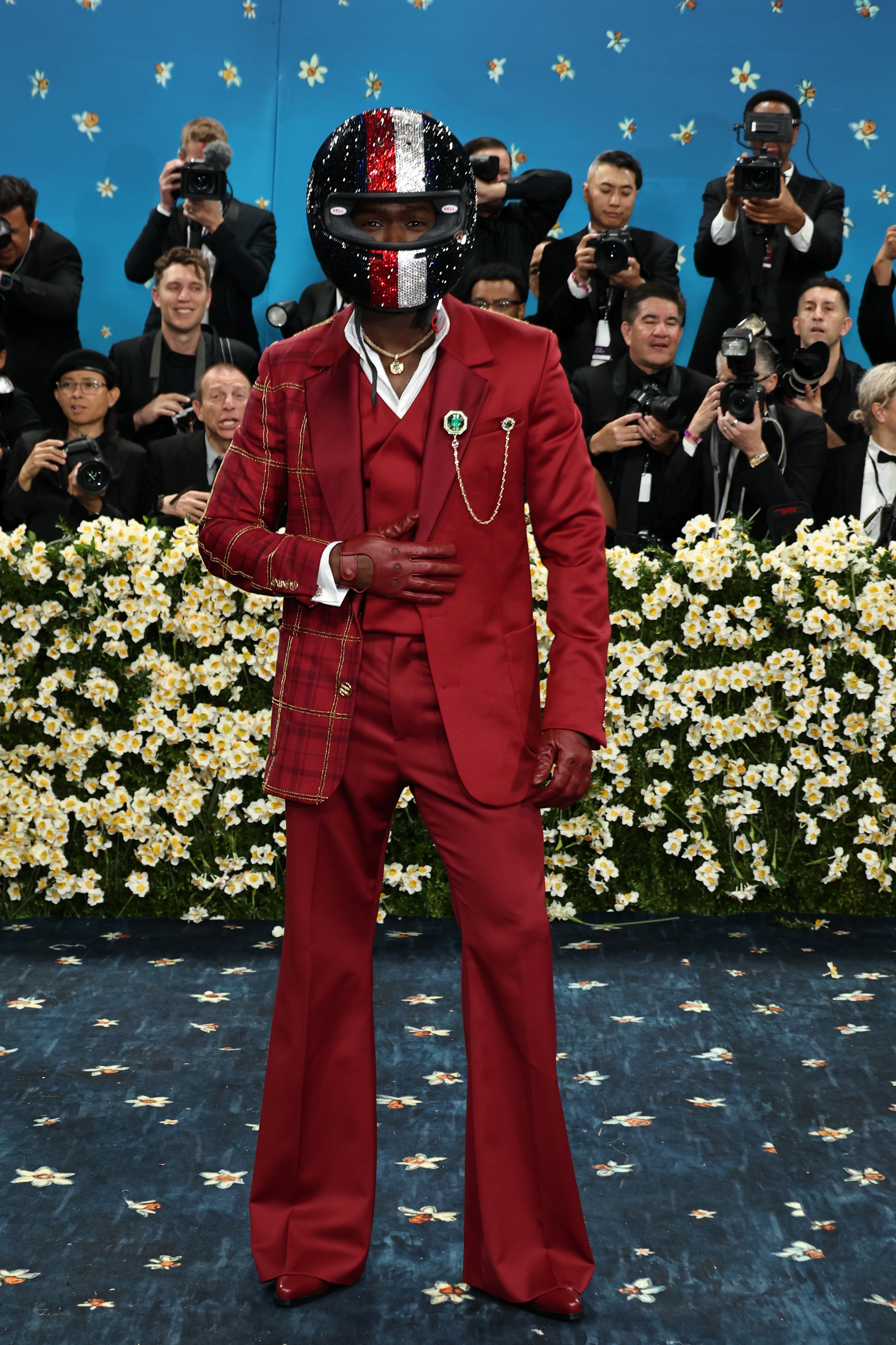 Person in a red plaid suit and striped helmet poses on blue floral-themed carpet, surrounded by photographers