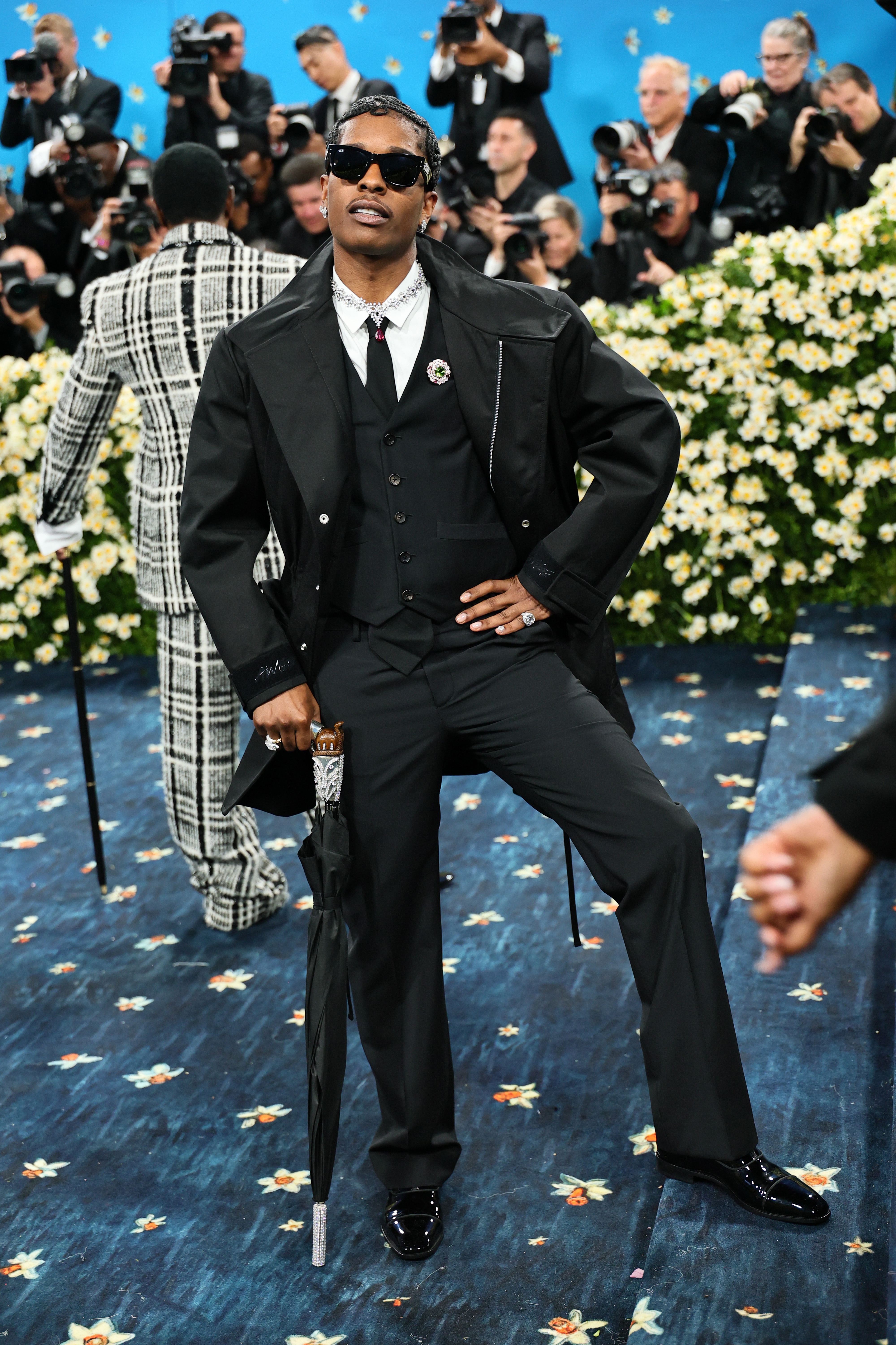 Person in a stylish three-piece suit with an umbrella on a red carpet, surrounded by photographers and floral arrangements