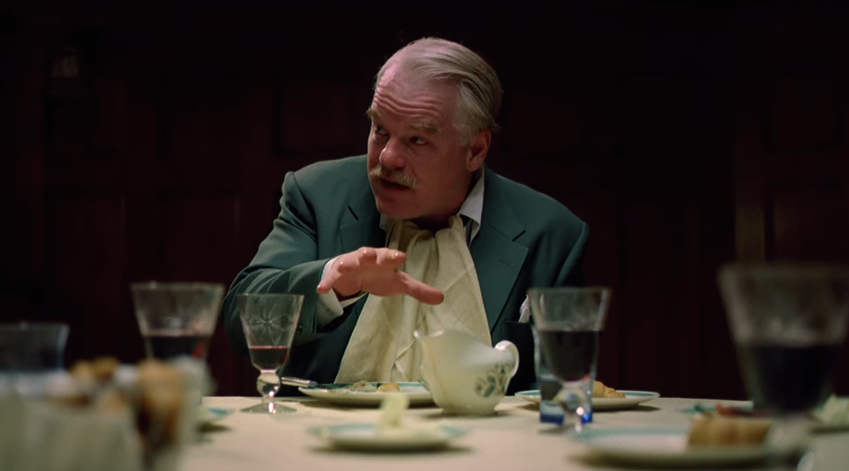 A person in a suit gestures at a dining table with glasses and plates, appearing to engage in conversation