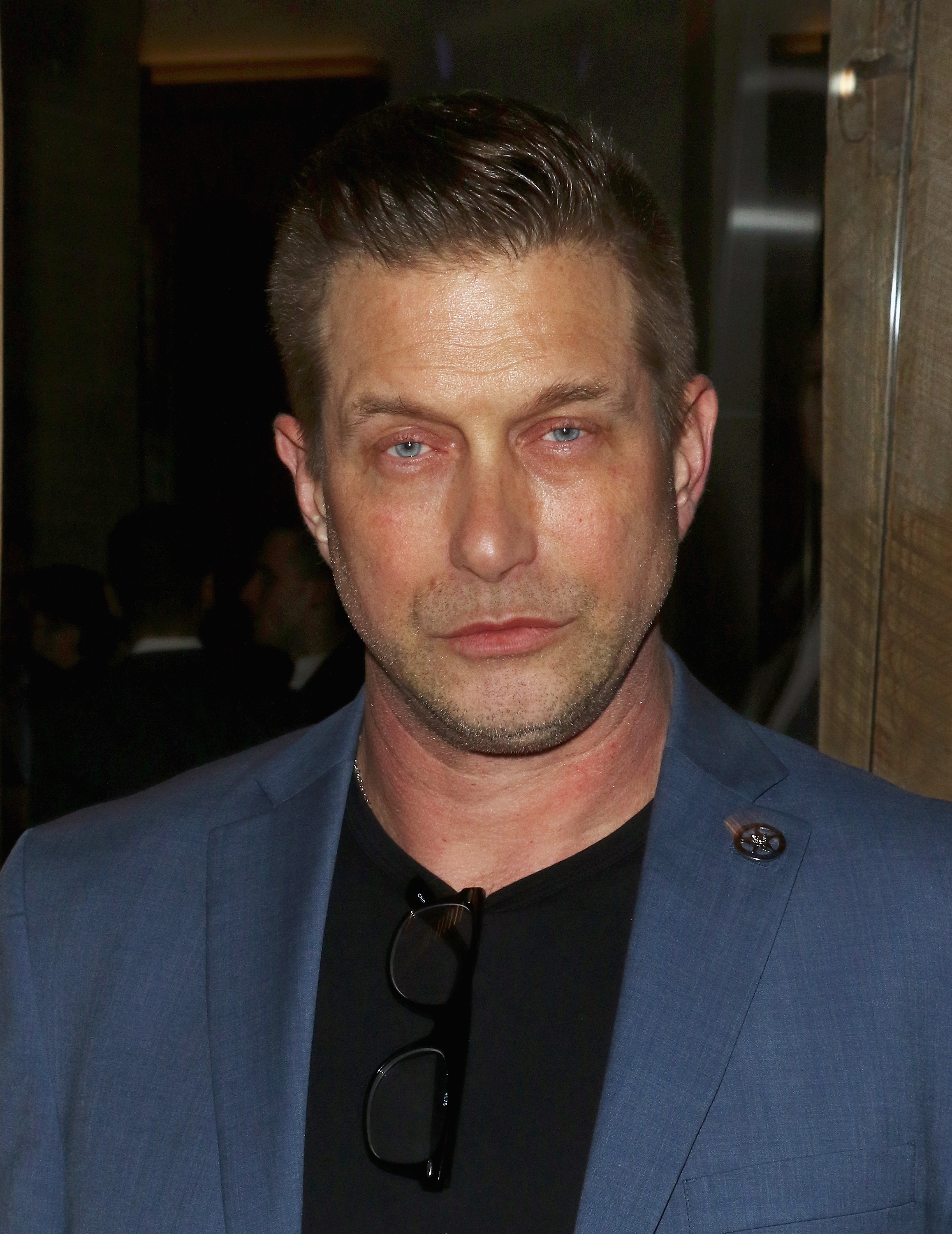 A person in a blue blazer and black shirt, standing at a formal event. They have short hair and are gazing directly at the camera