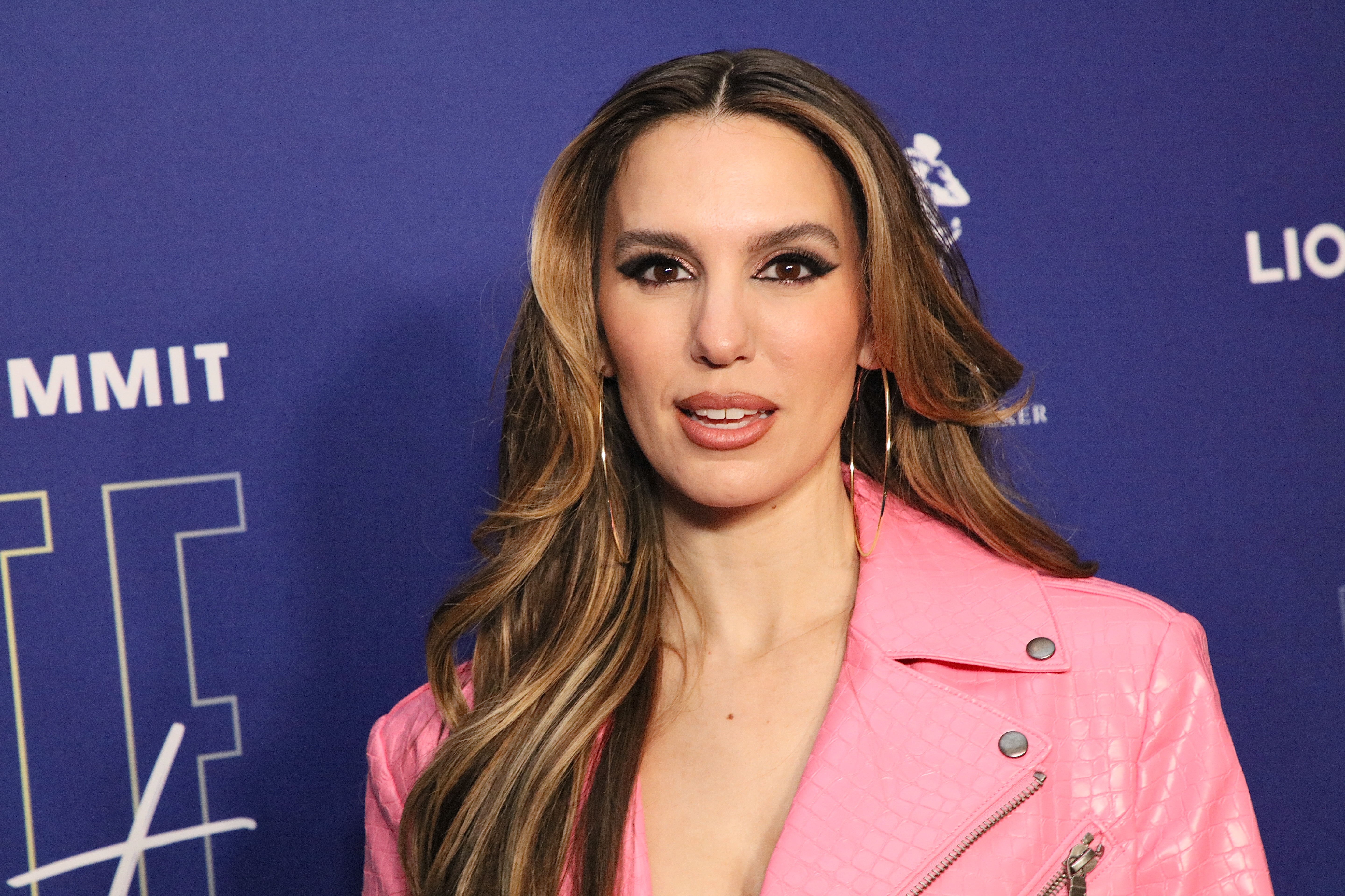 A person poses on a blue carpet wearing a stylish pink jacket