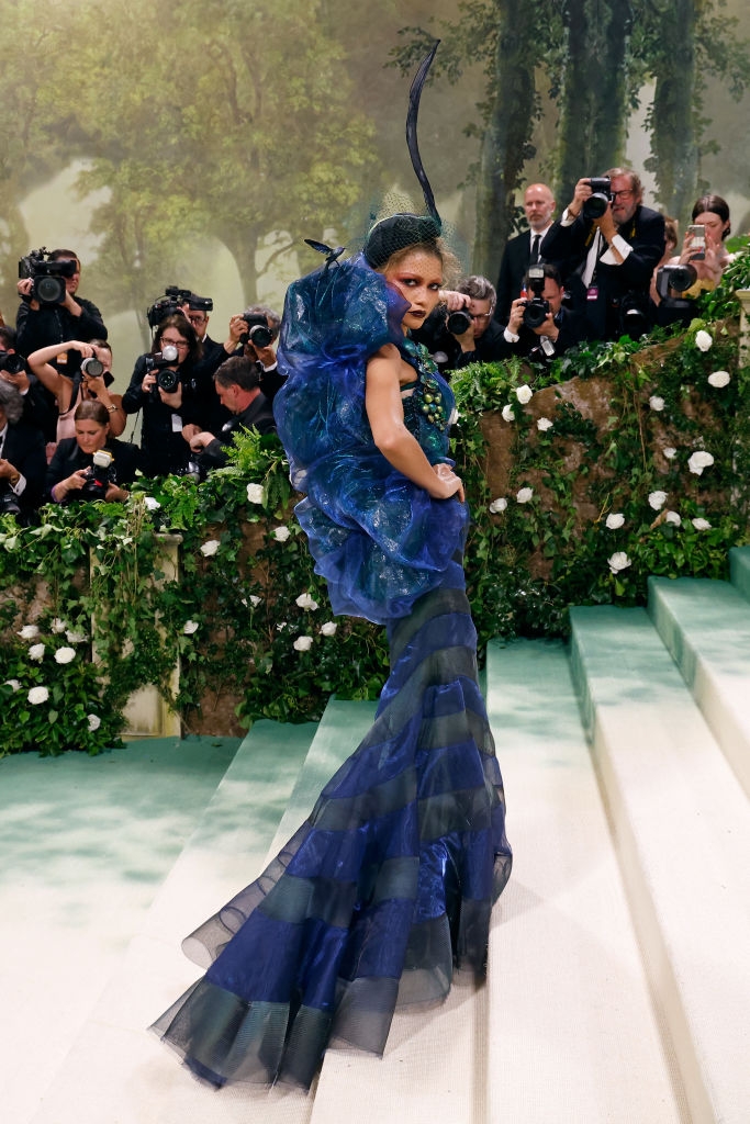 A person poses dynamically on steps in a sculptural, avant-garde gown with voluminous details, surrounded by photographers at an event