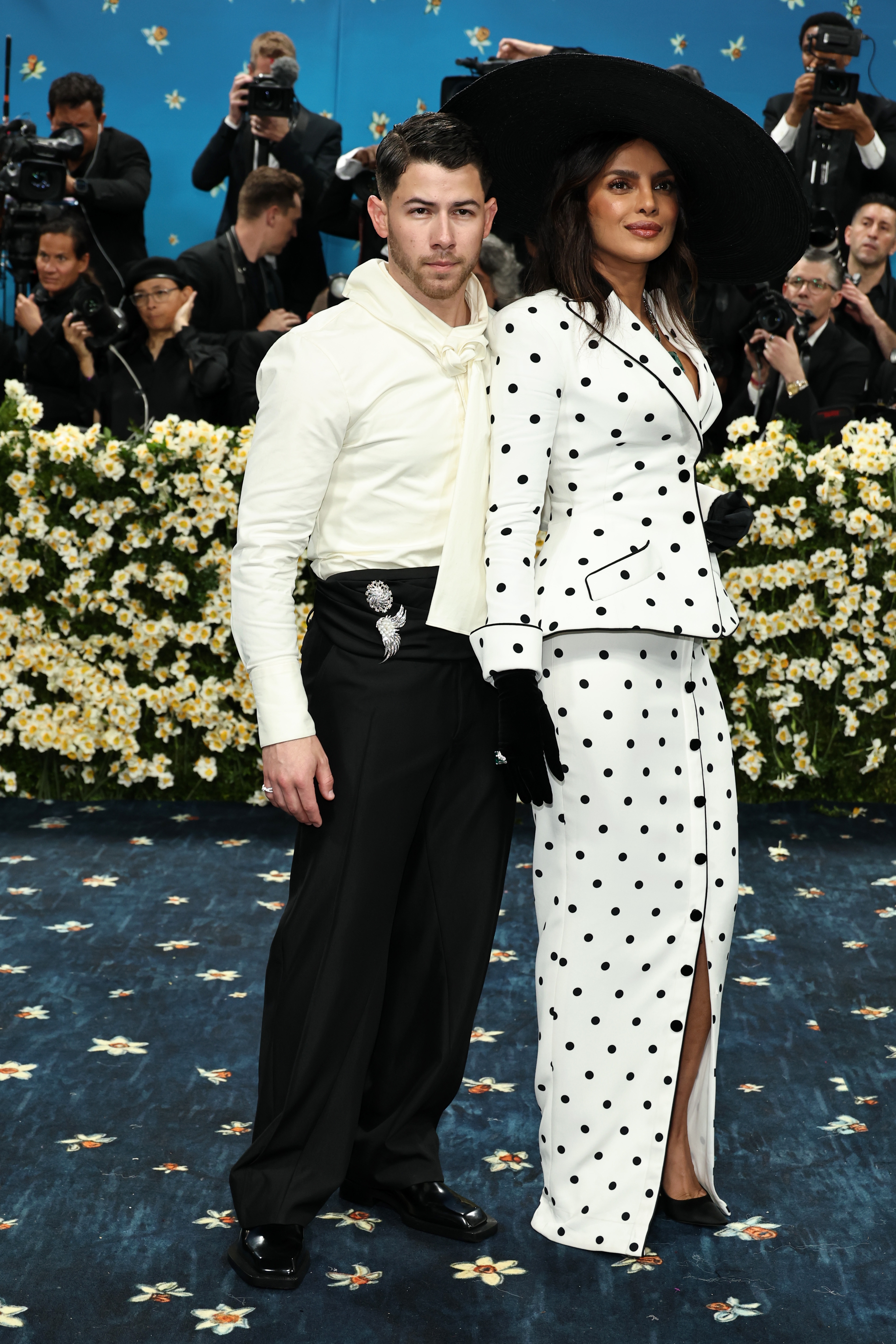 A man and woman pose on a floral-themed red carpet. The woman wears a polka dot suit with a wide-brim hat, while the man wears a tailored outfit
