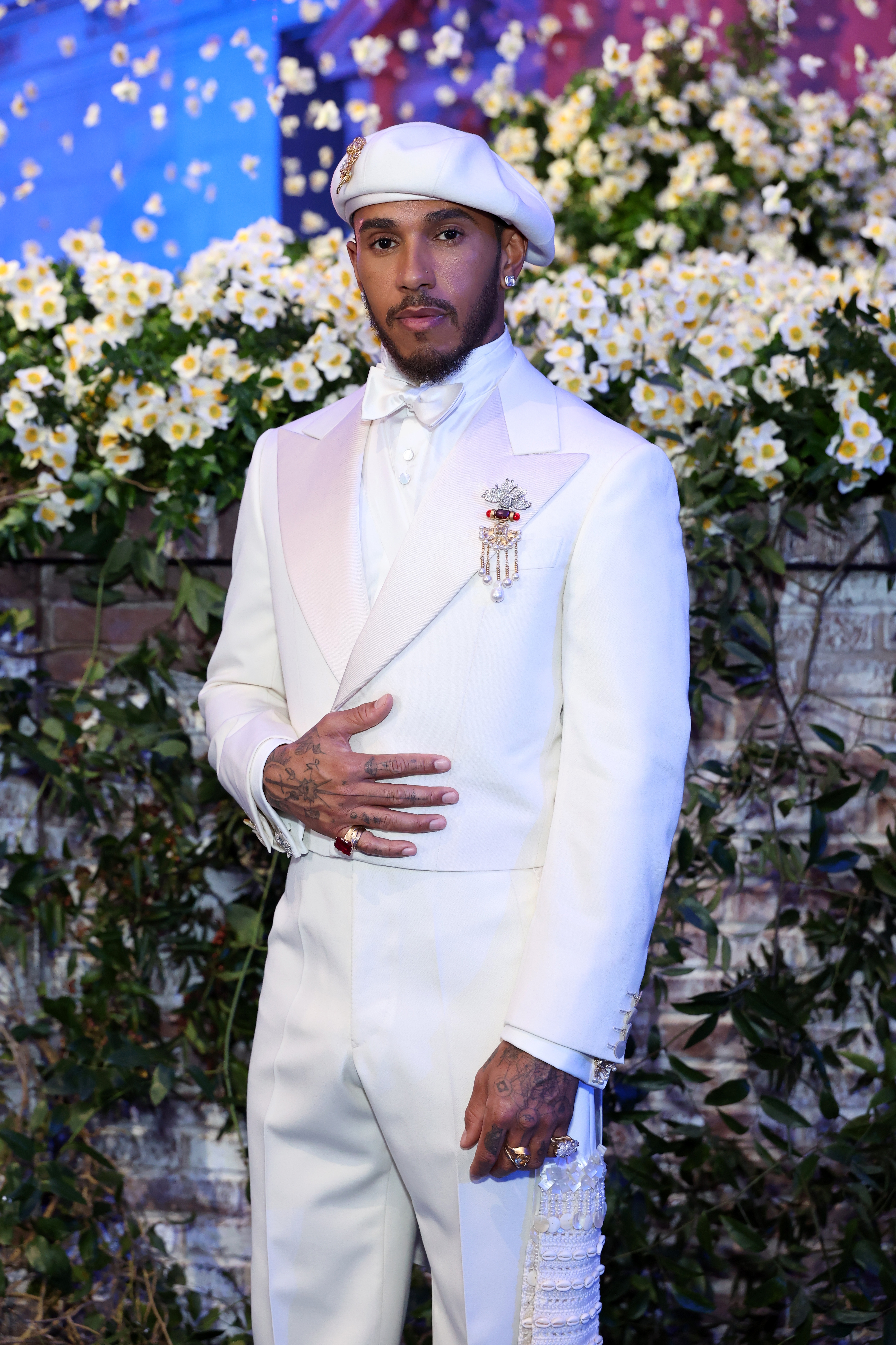 A person stands on a floral-decorated set in an elegant white suit with a beret and accessories, posing for the camera