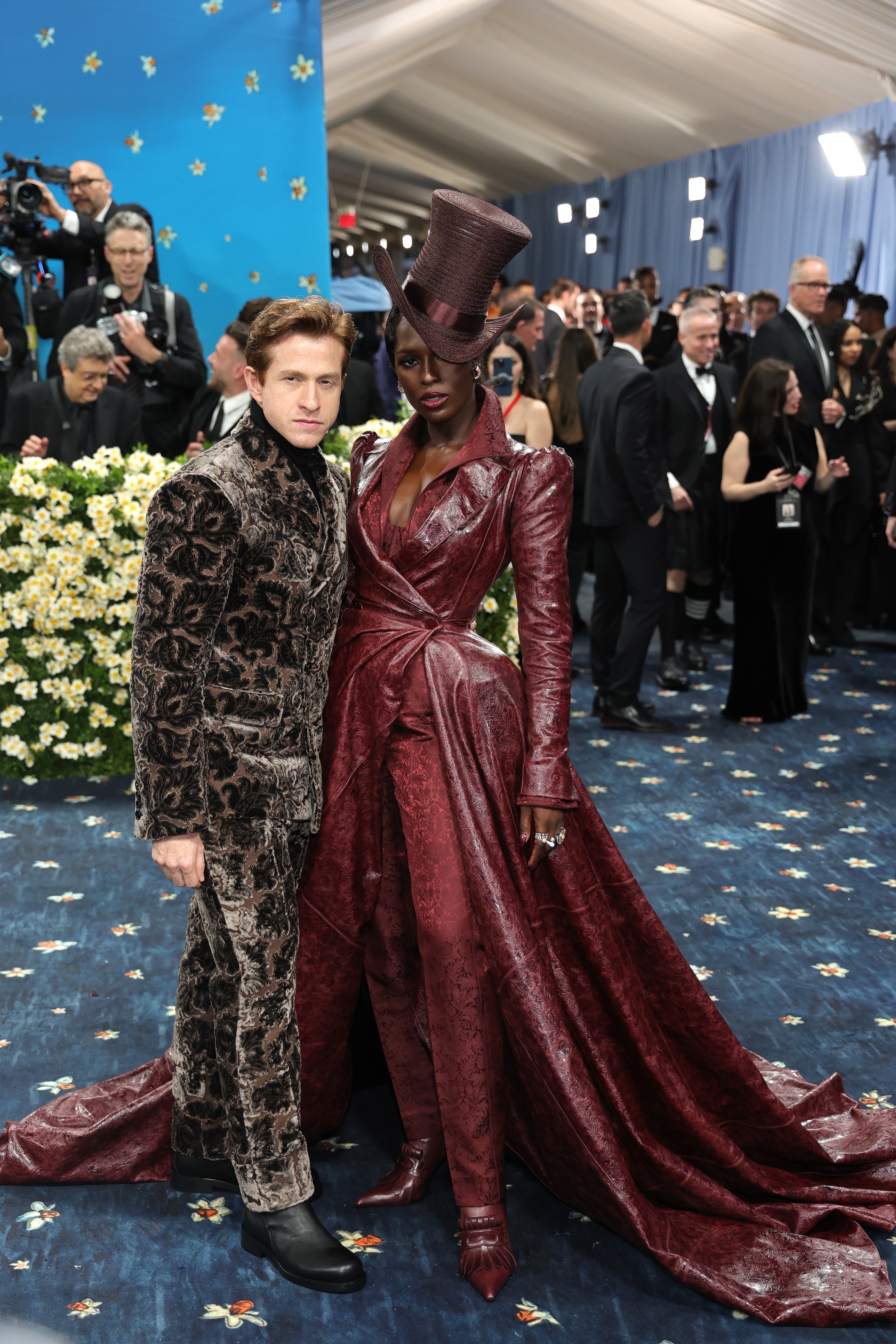 Two individuals on a red carpet, one in a velvet suit and the other in a striking textured gown with a dramatic hat, posed for photos