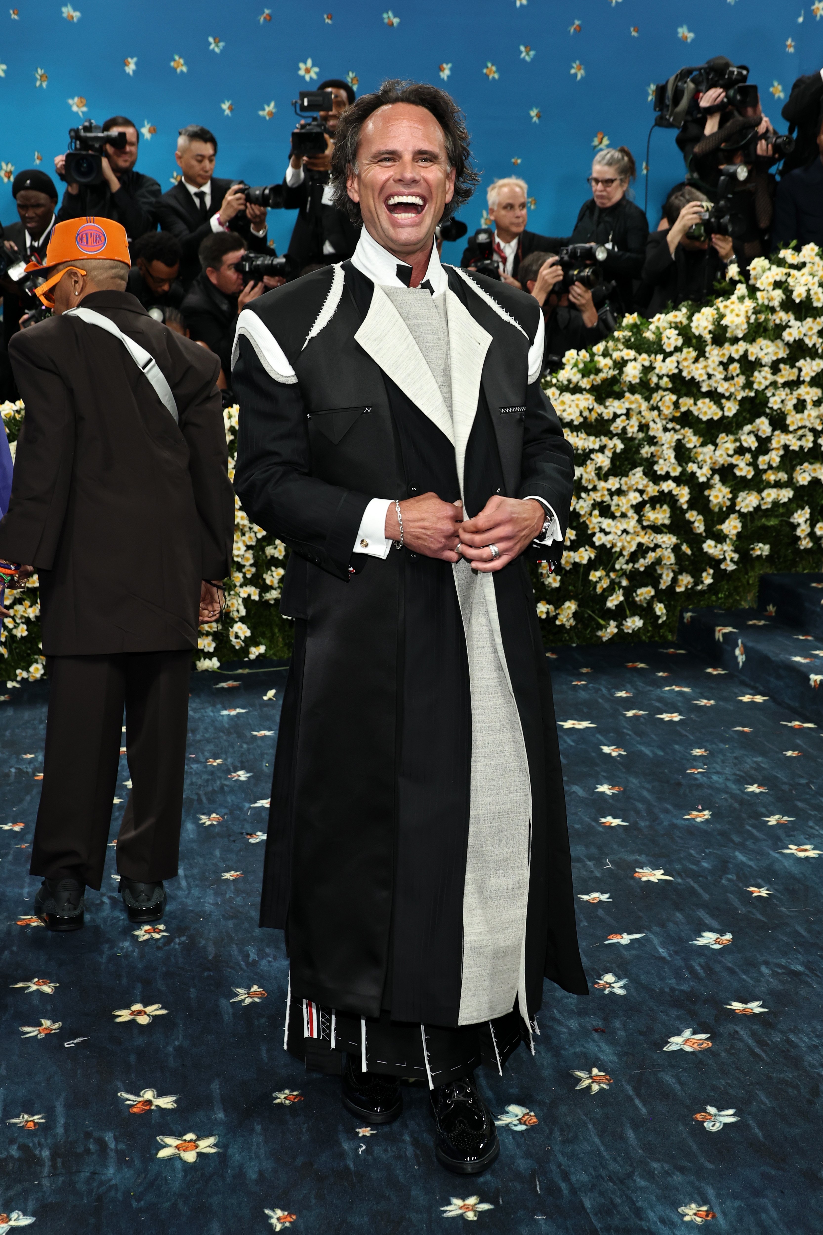 Person in a long, black and white suit with bold lapels, smiling on a floral-patterned carpet at a celebrity event. Photographers in the background