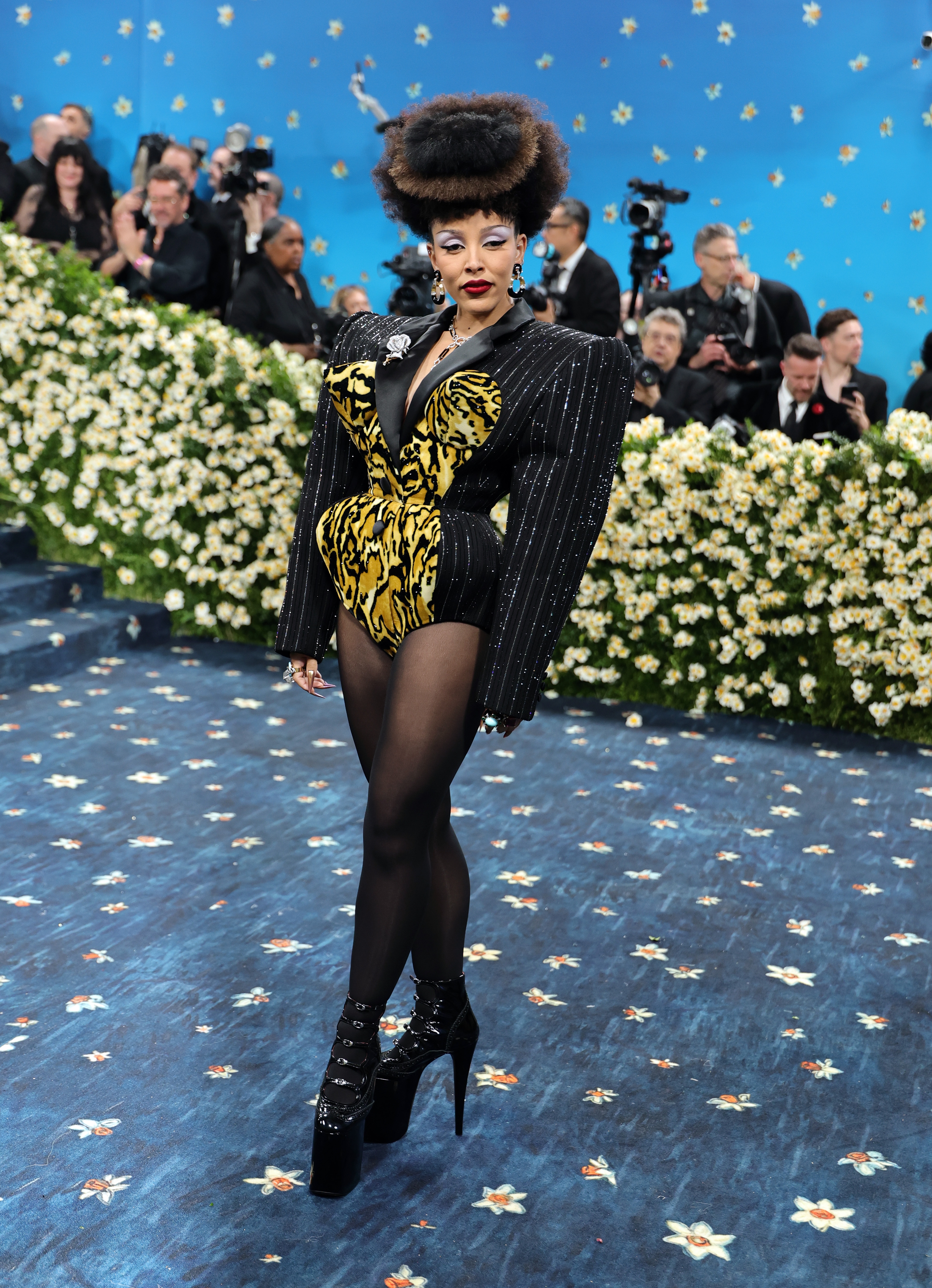 Celebrity in a bold, avant-garde outfit with structured black and gold elements, striking a pose on a floral-themed red carpet