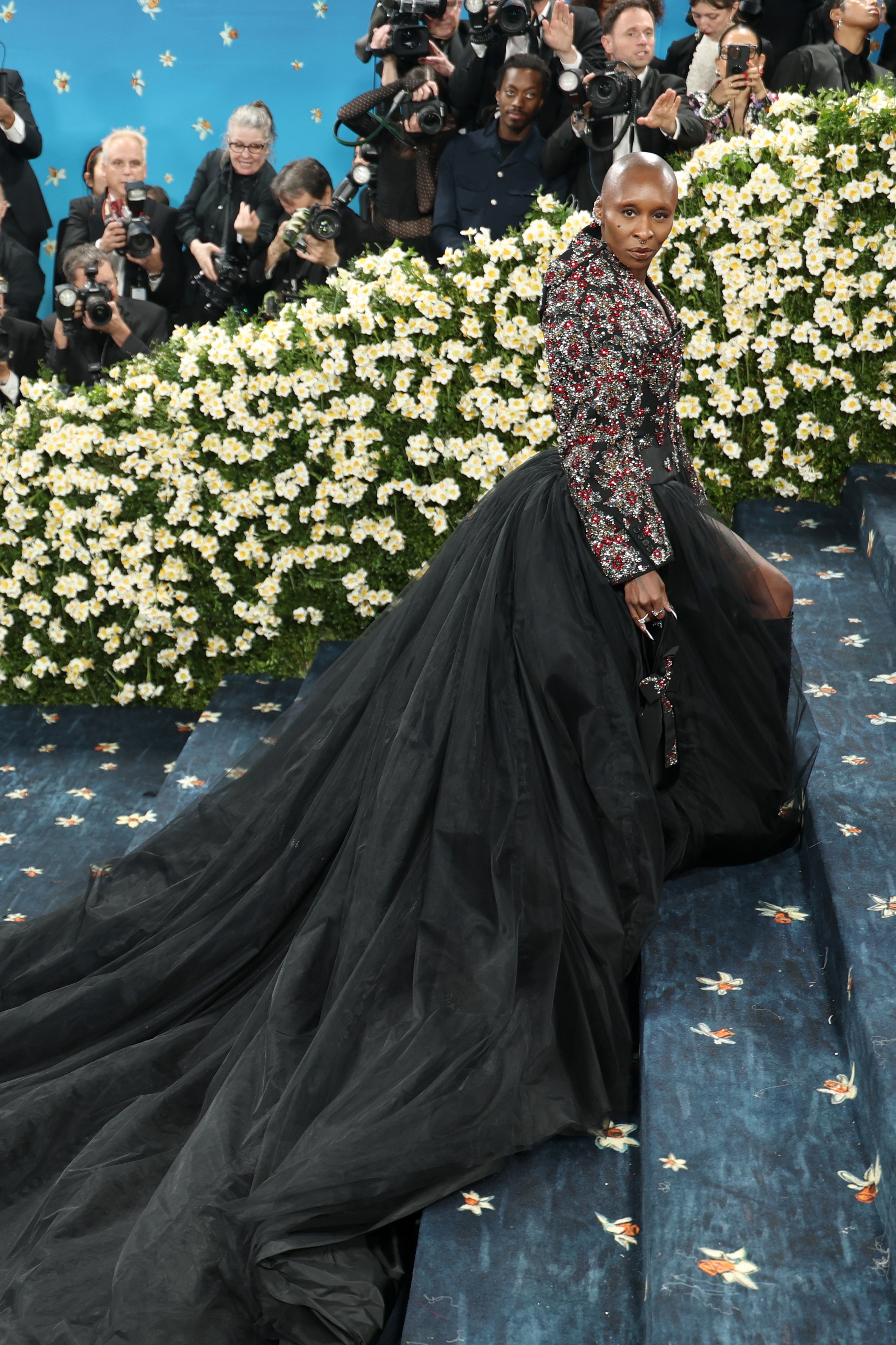 Person in ornate jacket with long black tulle train poses on blue carpeted steps, surrounded by photographers and floral backdrop