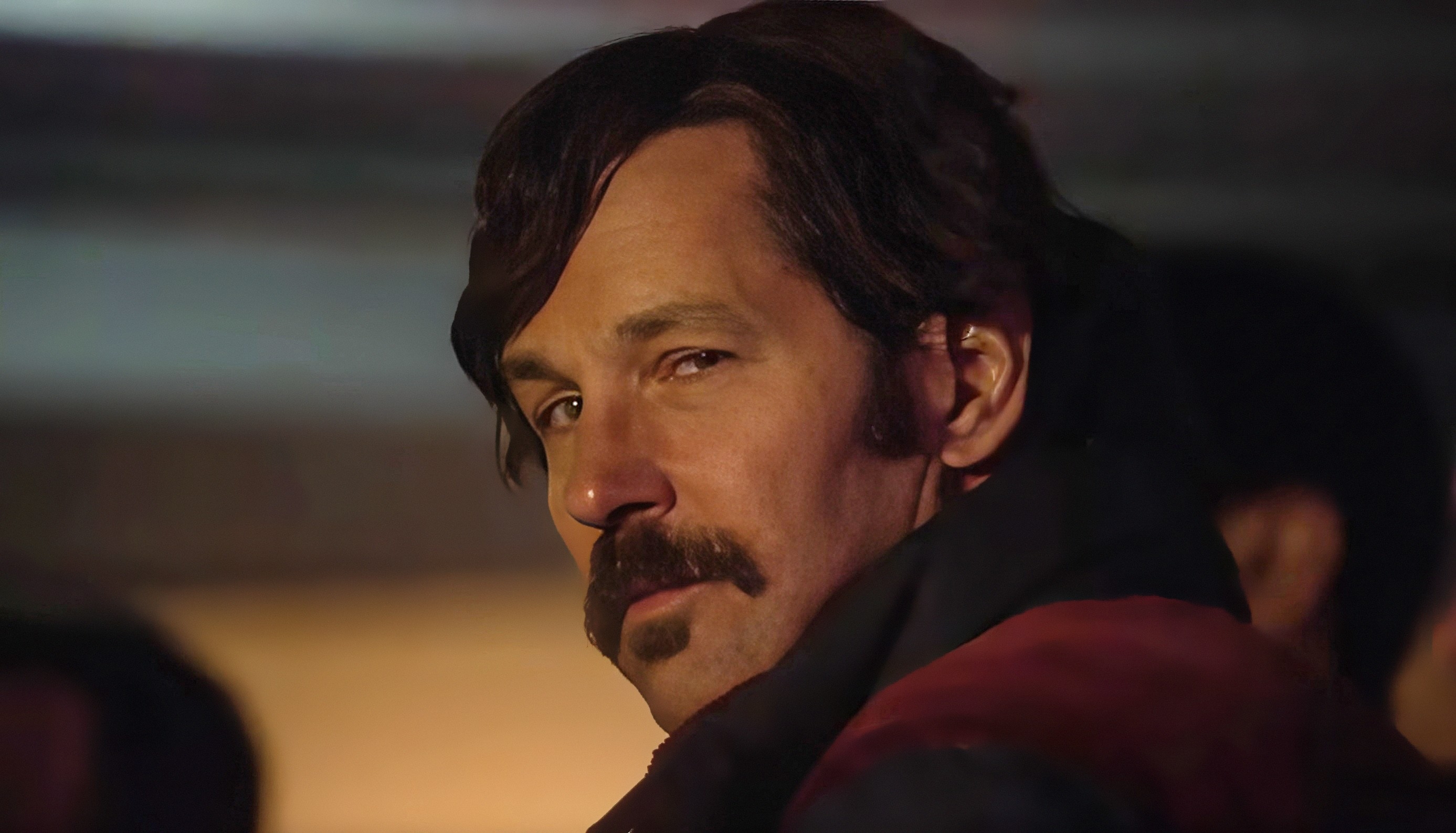 Man with a mustache and medium-length hair looking over his shoulder with a slight smile, appearing in a pensive moment