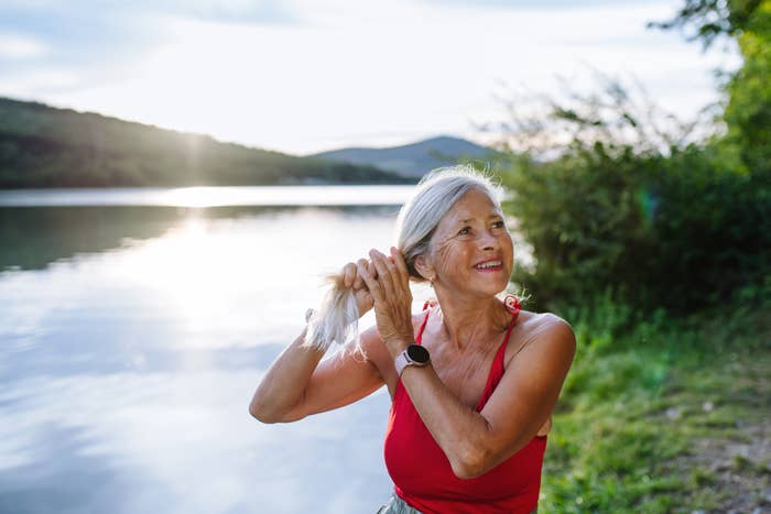 A woman in a sleeveless top stands by a lake, smiling and styling her hair, with trees and hills in the background