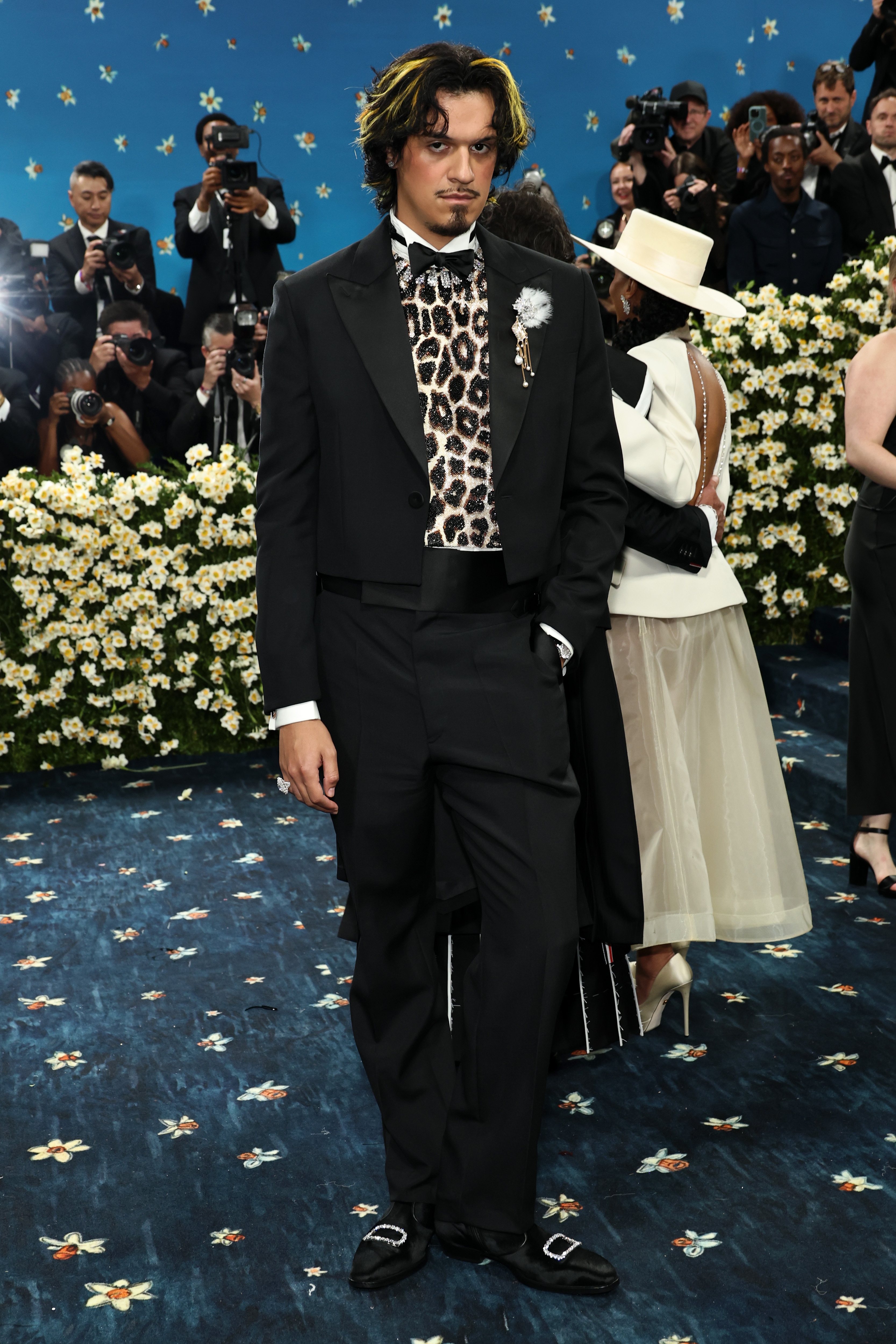 A person in a tuxedo with a leopard-print shirt on a floral-patterned carpet, surrounded by photographers at a formal event