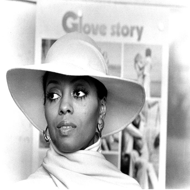 Person wearing a wide-brim hat and scarf in front of a "Glove Story" poster, looking to the side. Black-and-white photo