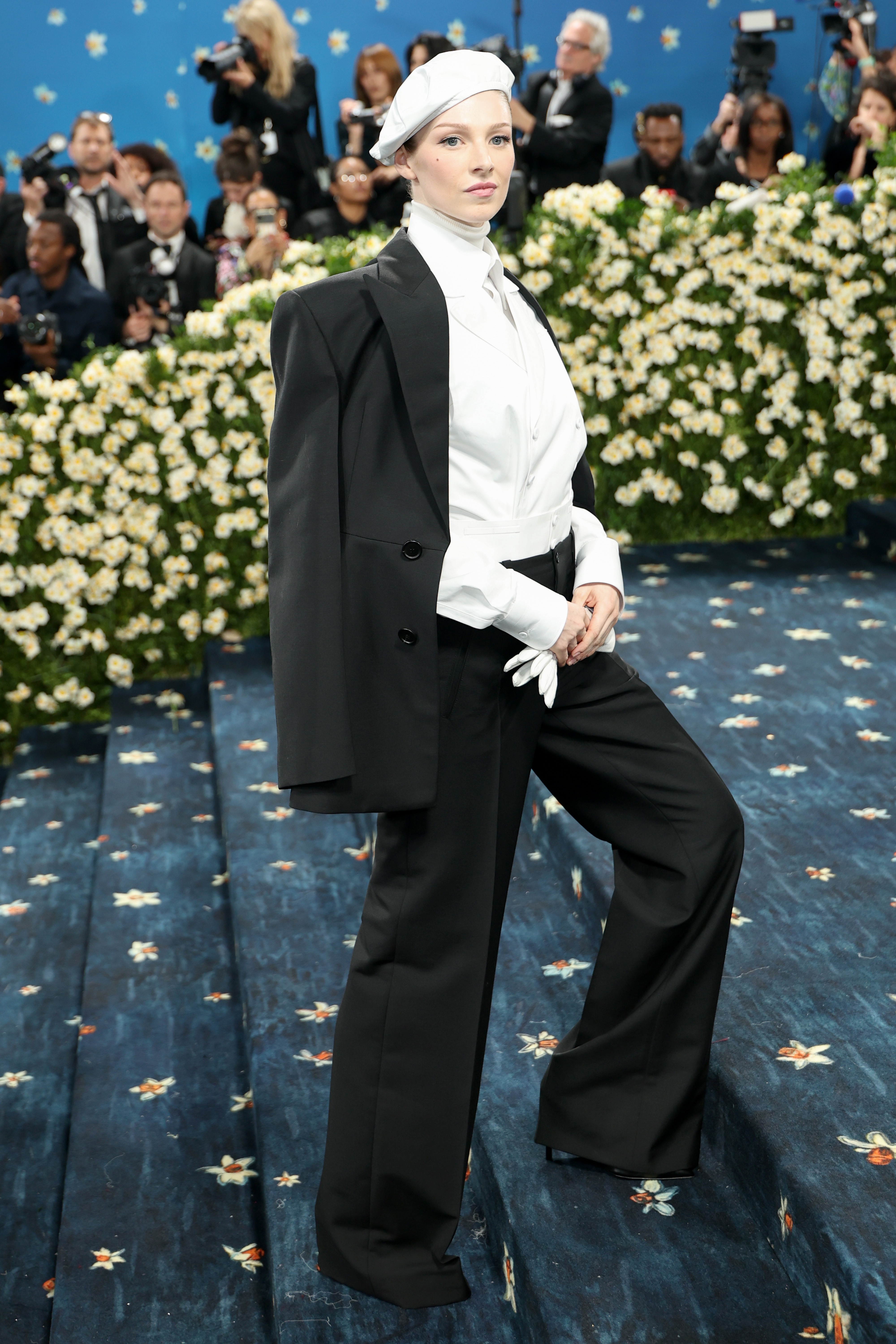 Person in a stylish black suit and hat poses confidently on a floral-themed red carpet event