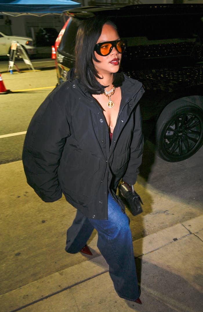 Person wearing a puffy jacket, flared jeans, and sunglasses, walking on a sidewalk at night