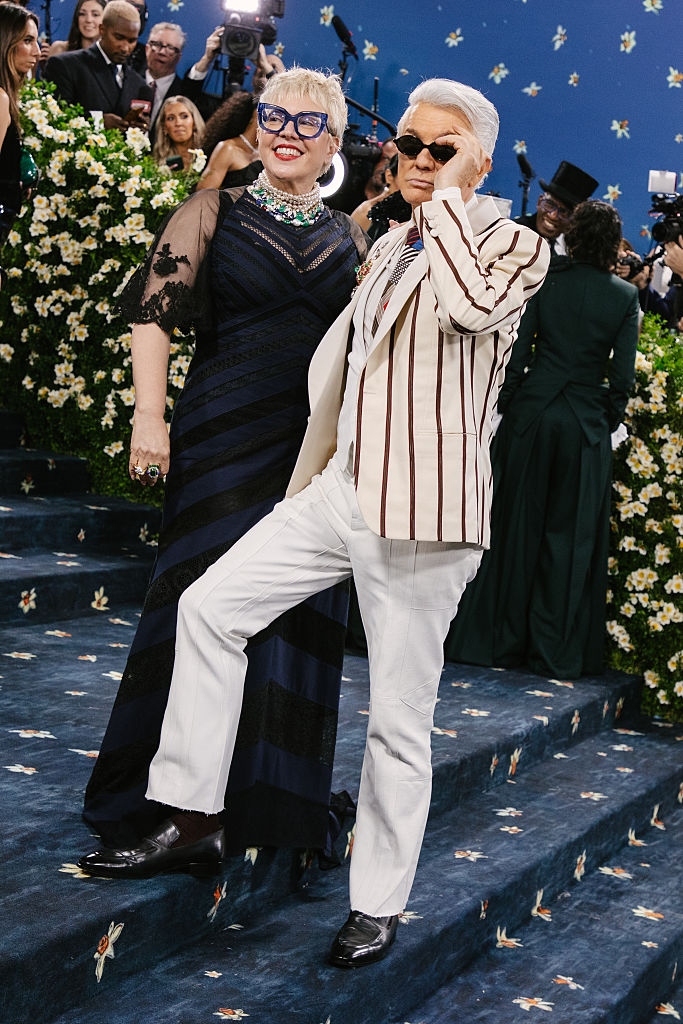 Catherine wears a dark gown with lace sleeves, Baz a striped blazer and solid pants with sunglasses