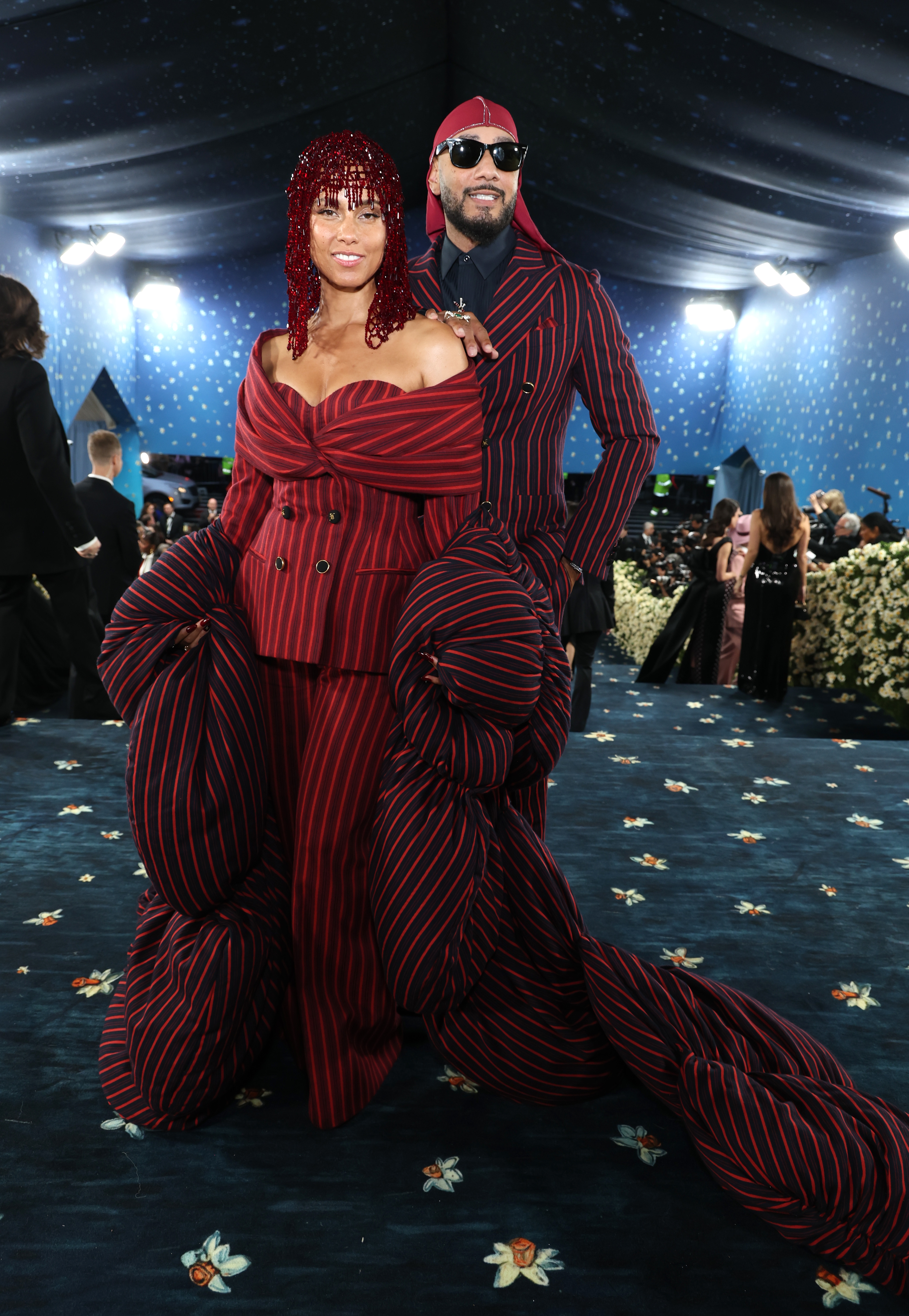 Two individuals wearing coordinated red and black striped outfits on a red carpet event, one in an off-shoulder dress, the other in a suit and sunglasses