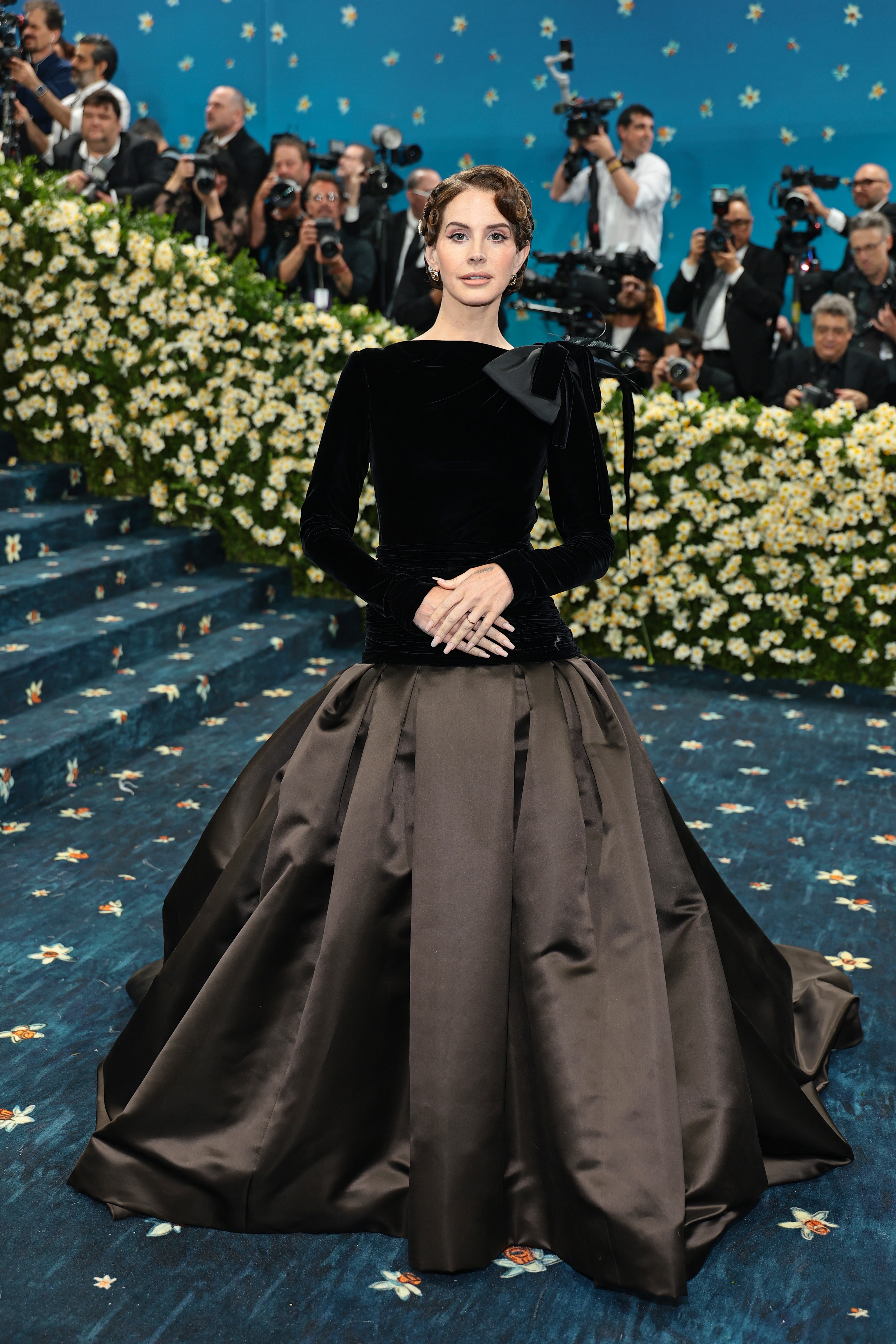 Person on an event carpet wearing an elegant floor-length gown with long sleeves and a voluminous skirt, surrounded by photographers