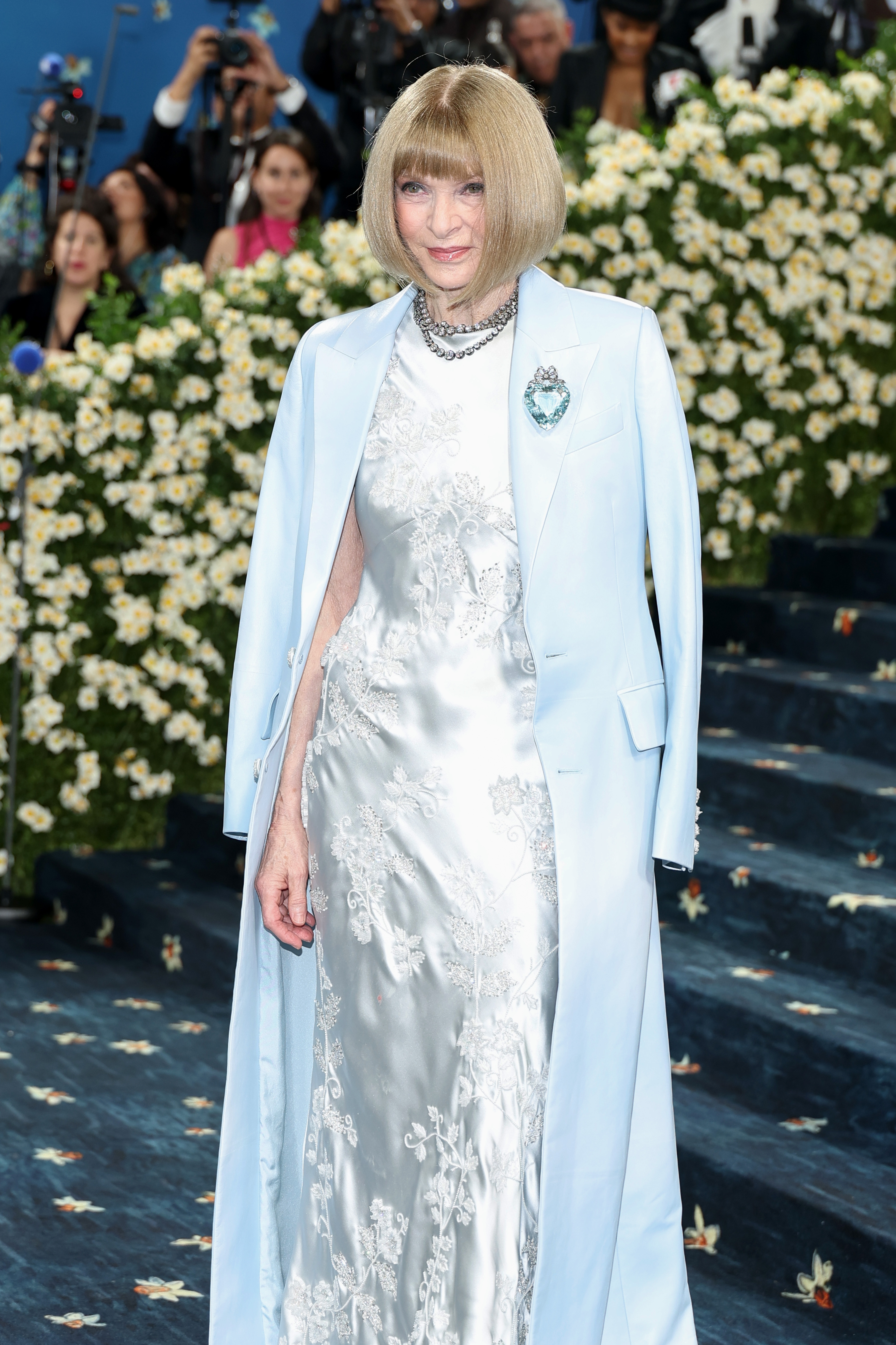 A person on a red carpet wears a long, elegant dress with floral patterns, paired with a matching coat. There are flowers in the background
