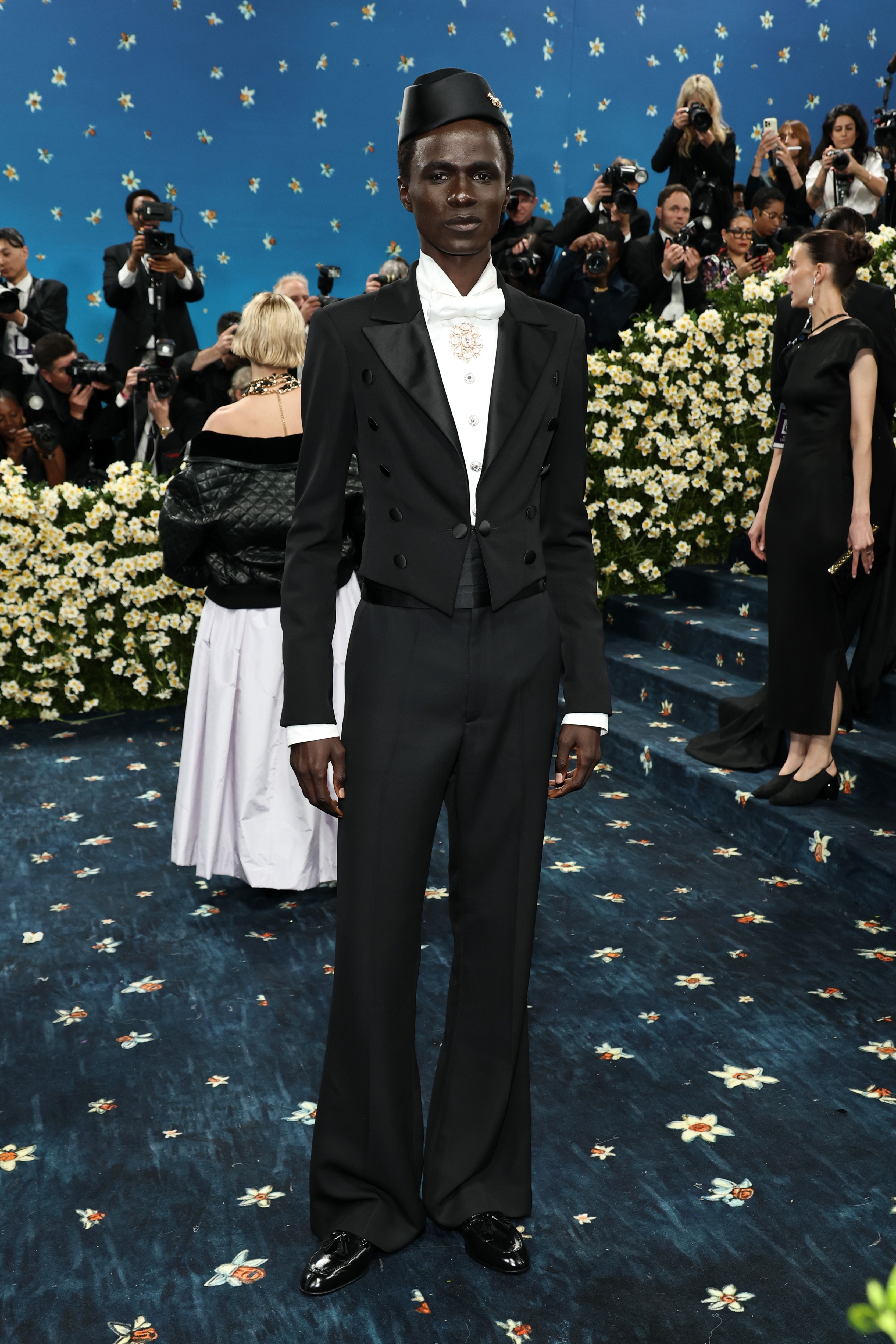 Person on patterned carpet wearing an elegant, tailored tuxedo with a high collar and decorative brooch, surrounded by flowers and photographers