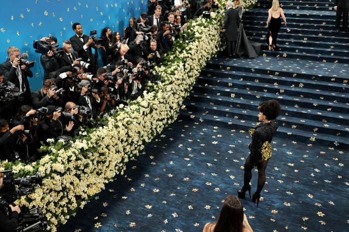 A person poses confidently on a floral-decorated staircase at a formal event as photographers capture the moment