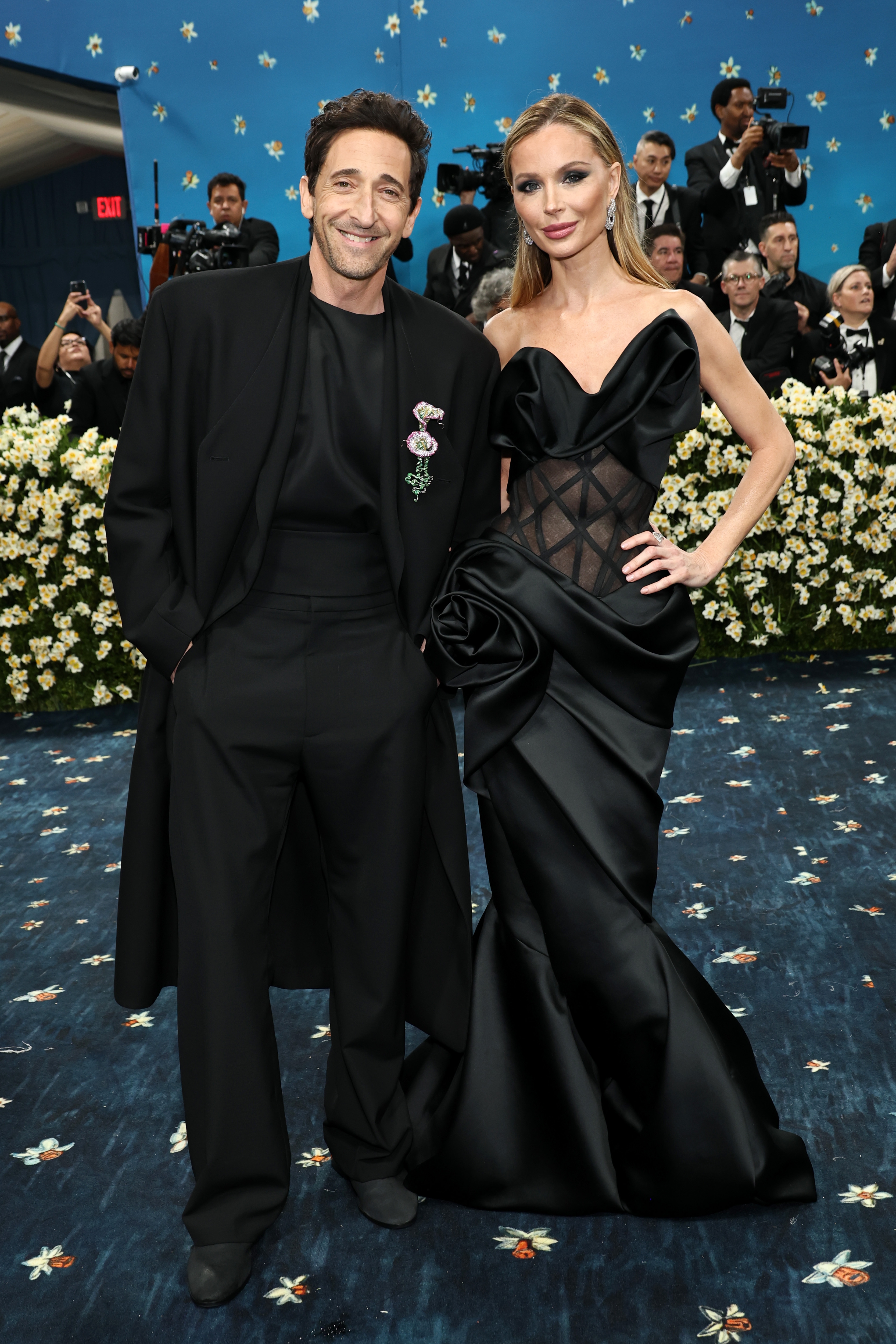 Two celebrities posing on a red carpet; the man in a dark suit and the woman in a black gown with sheer details and floral embellishments