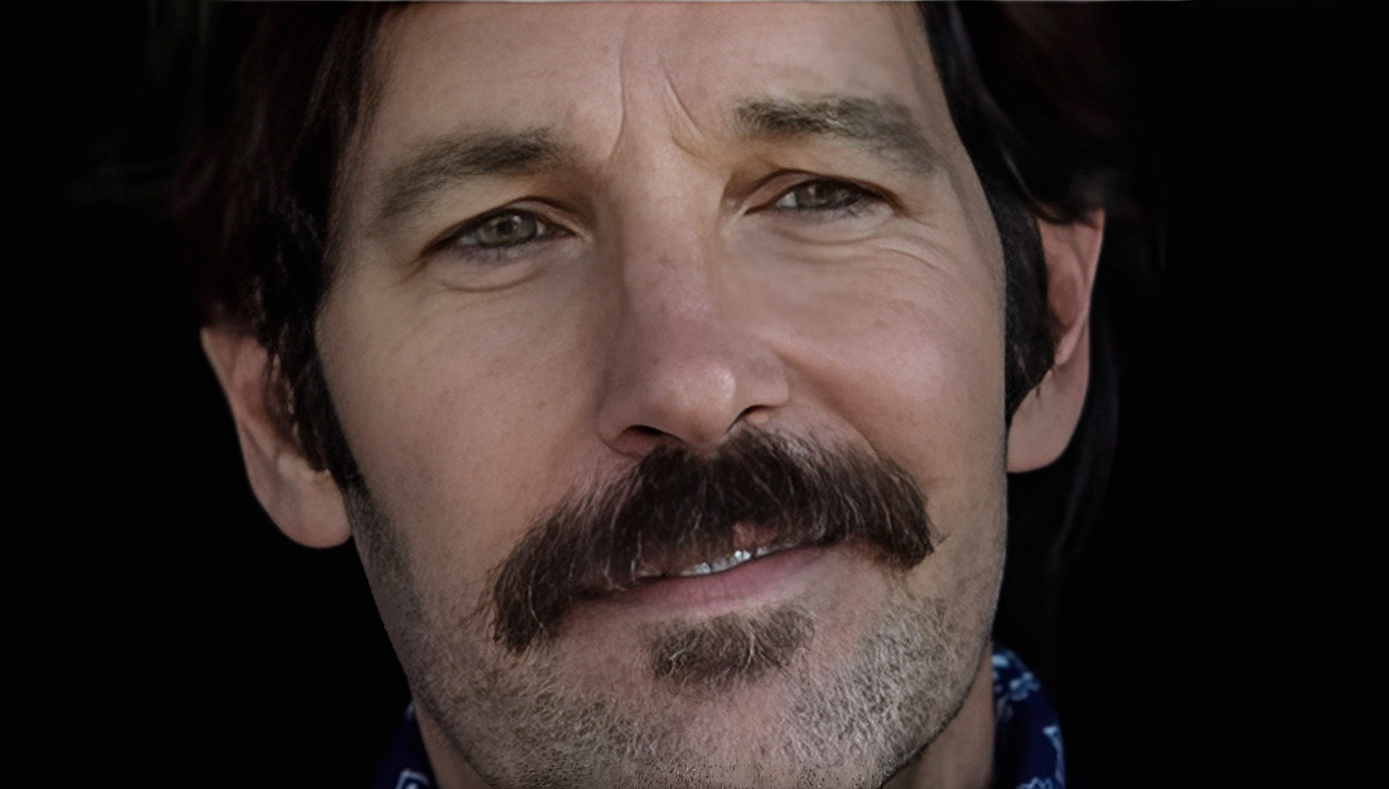 A person with short hair and a mustache looks slightly away from the camera, wearing a bandana around their neck