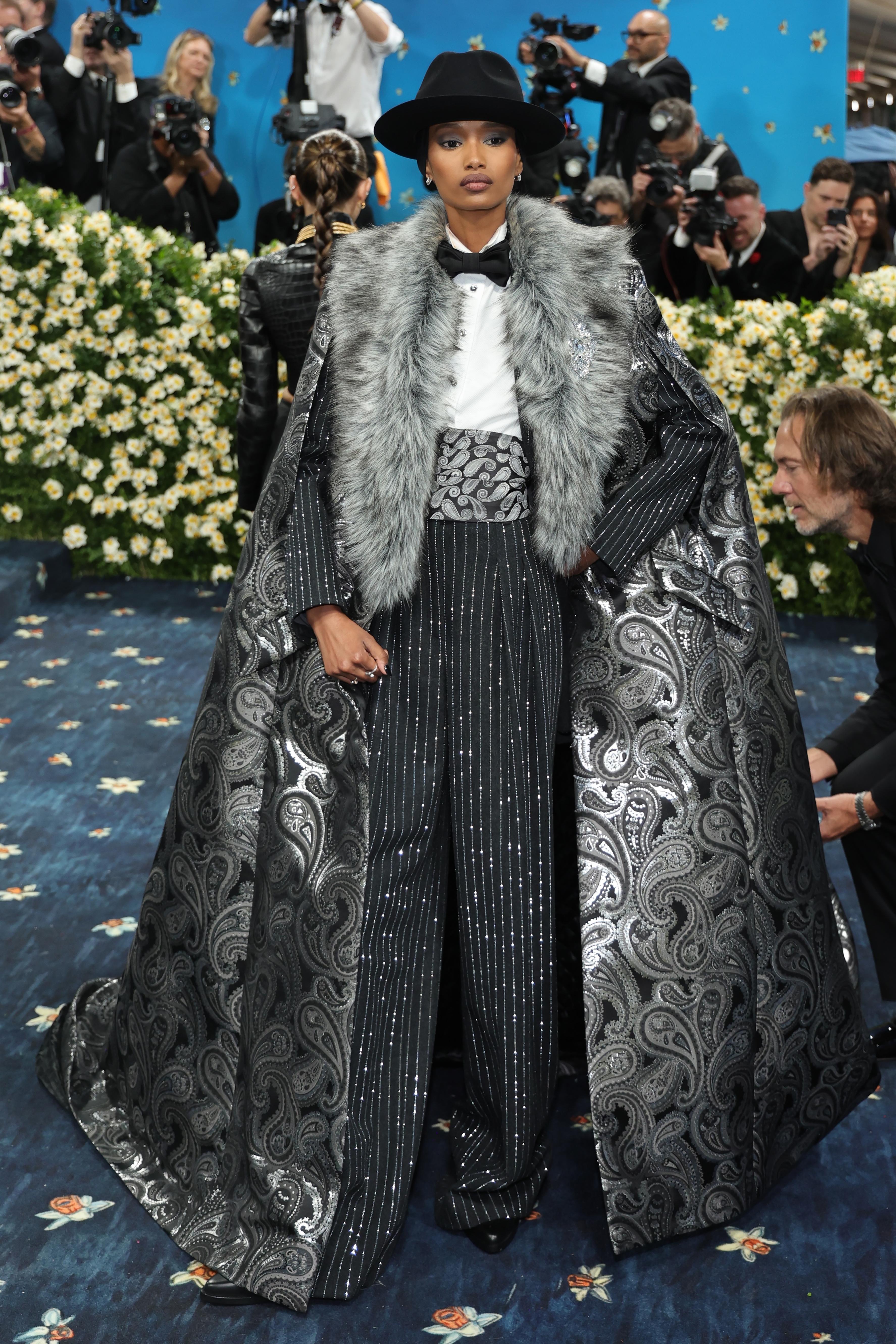 Person in a wide-brim hat and embroidered suit with a silver fur cape on a red carpet, surrounded by photographers and floral backdrop