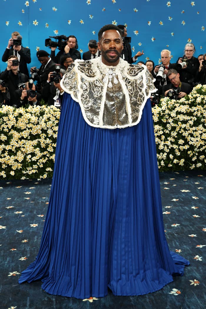 Person in elaborate cape with sequined top poses for photographers at an event, in front of floral backdrop